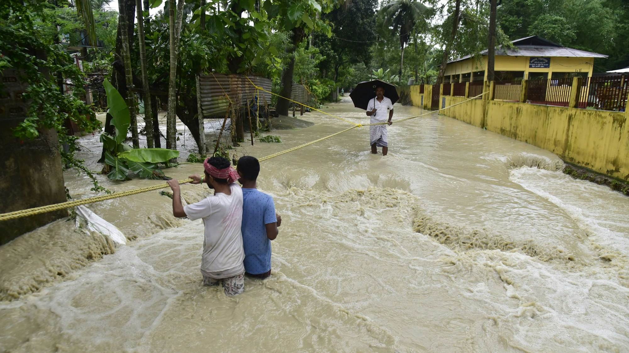 21.06.2023, Indien, Guwahati: Menschen halten sich an einem Führungsseil fest, als sie durch das Hochwasser einer überschwemmten Straße waten.