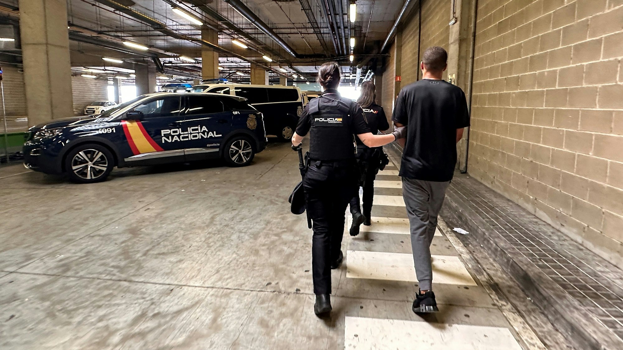 Ein Mann wird von der spanischen Polizei verhaftet abgeführt. Die auf dem Foto sichtbaren Menschen haben der Kamera den Rücken zugedreht. Im Hintergrund befinden sich Polizeiwagen.
