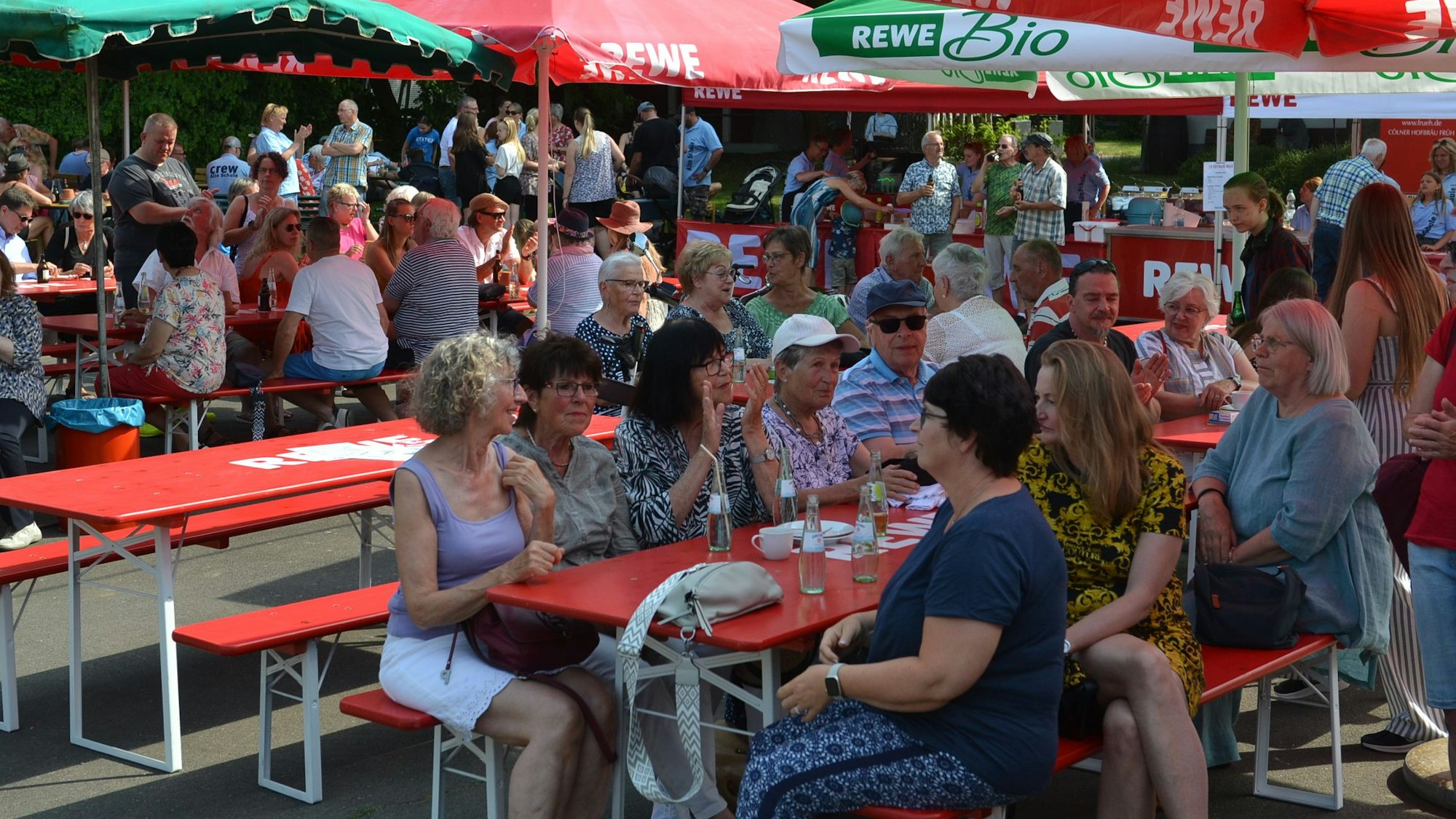 Besucher eines Dorffests sitzen an Tischen und Bänken unter aufgestellten Sonnenschirmen.