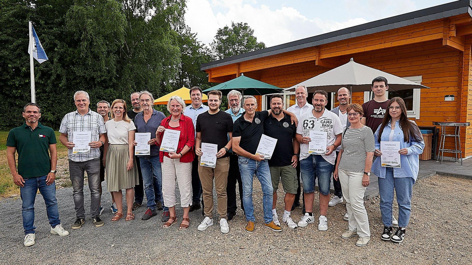 Eine Gruppe von Männern und Frauen halten Förderbescheide in der Hand. Sie stehen vor einem Holzhaus.