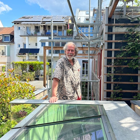 Ein Mann mit grauem Haar und Bart steht im kurzärmeligen Hemd auf einer Dachterrasse.
