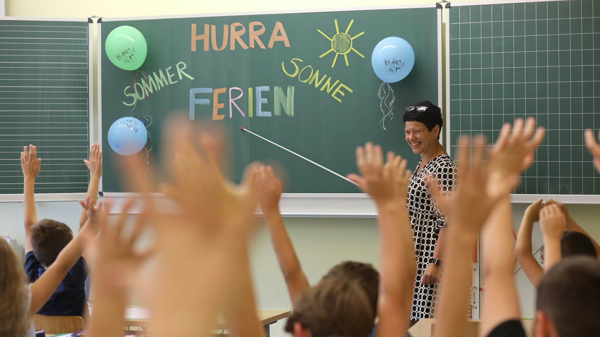 „Hurra Sommerferien“ steht an einer Tafel in der zweiten Klasse einer Grundschule. Eine Lehrerin zeigt mit einem Zeigestock lächelnd auf die Worte, die Kinder heben jubelnd ihre Arme.