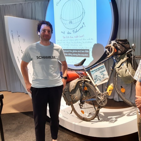 Das Bild zeigt zwei Herren vor einem als Ausstellungsstück aufgestellten alten Fahrrad mit Satteltaschen.