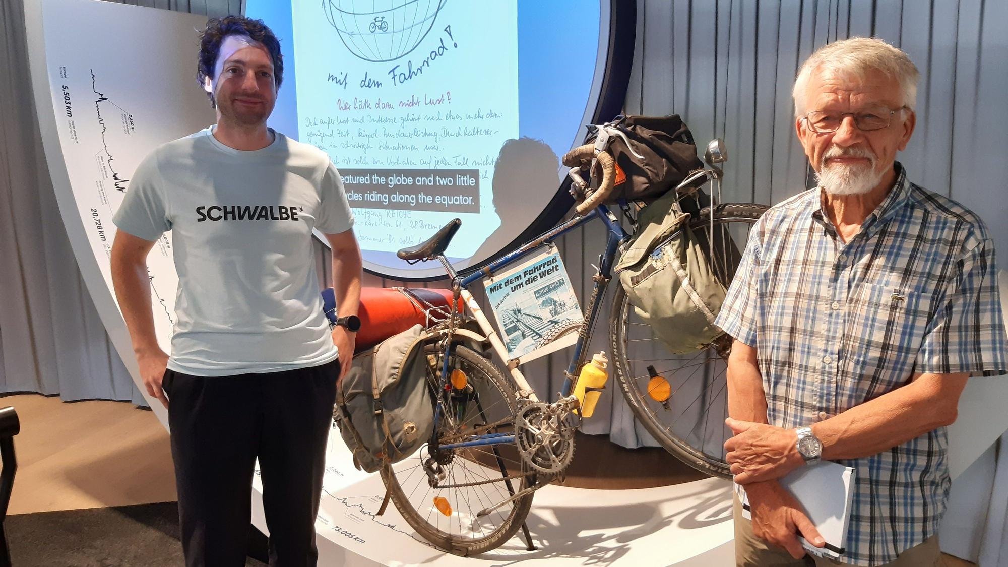 Das Bild zeigt zwei Herren vor einem als Ausstellungsstück aufgestellten alten Fahrrad mit Satteltaschen.