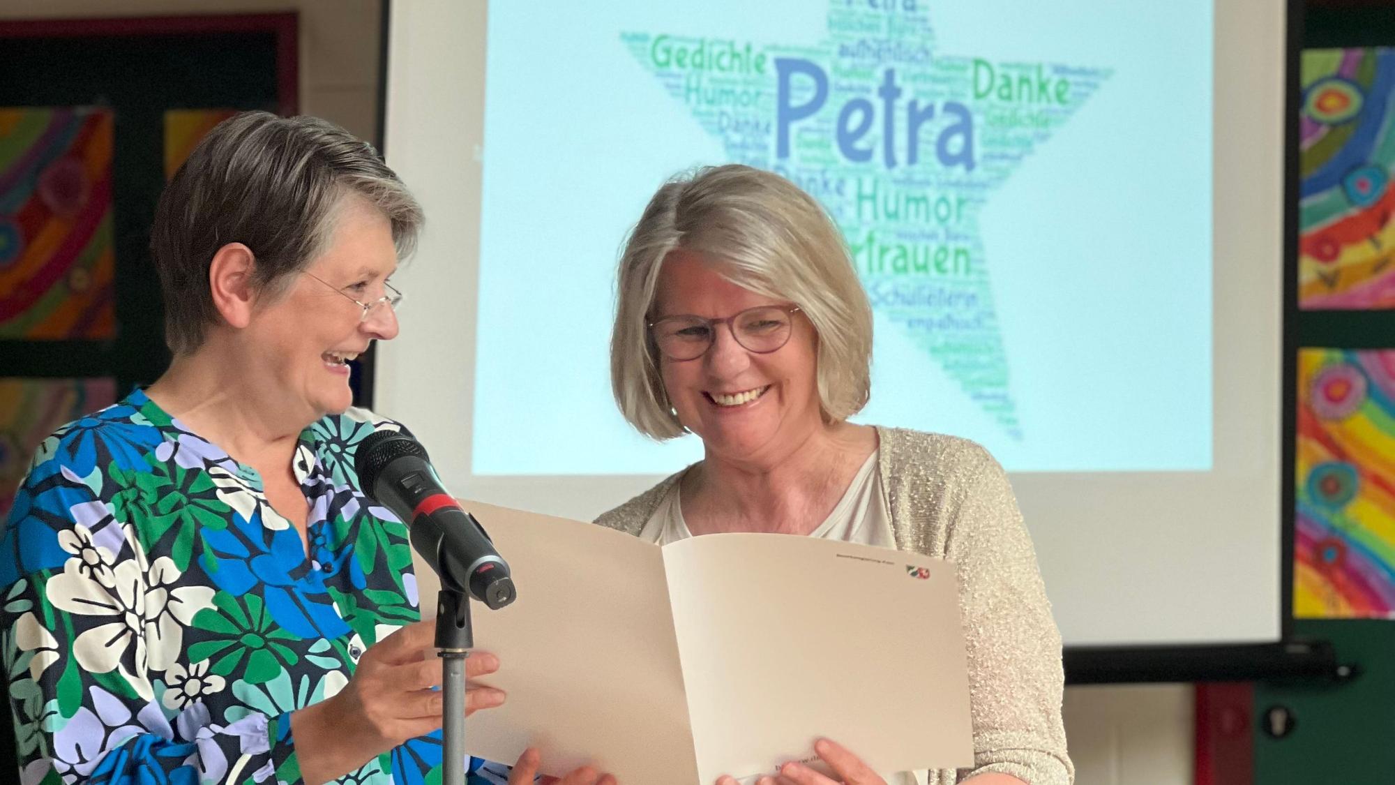 Es sind die beiden Frauen auf einer Bühne zu sehen. Im Hintergrund steht eine Leinwand, auf die ein Stern mit dem Namen „Petra“ projiziert wird.