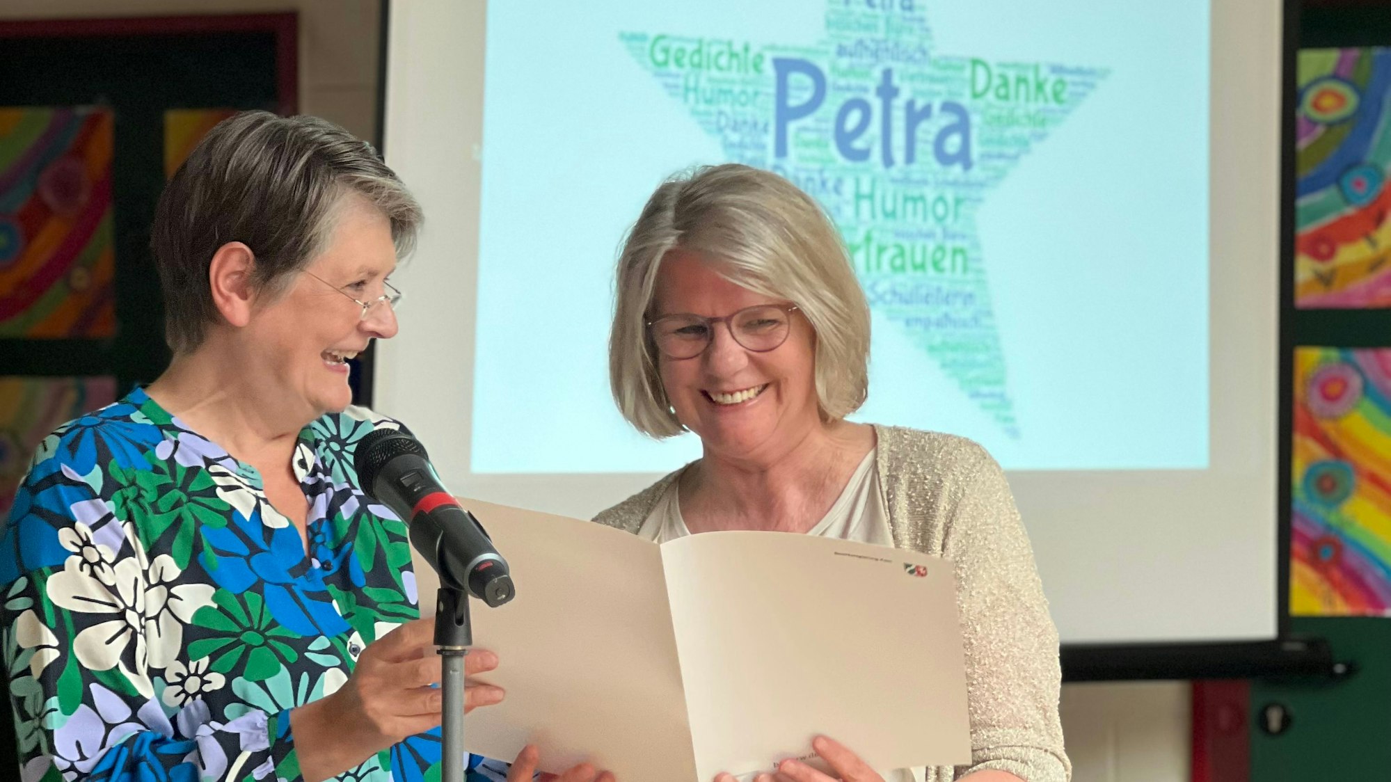 Es sind die beiden Frauen auf einer Bühne zu sehen. Im Hintergrund steht eine Leinwand, auf die ein Stern mit dem Namen „Petra“ projiziert wird.