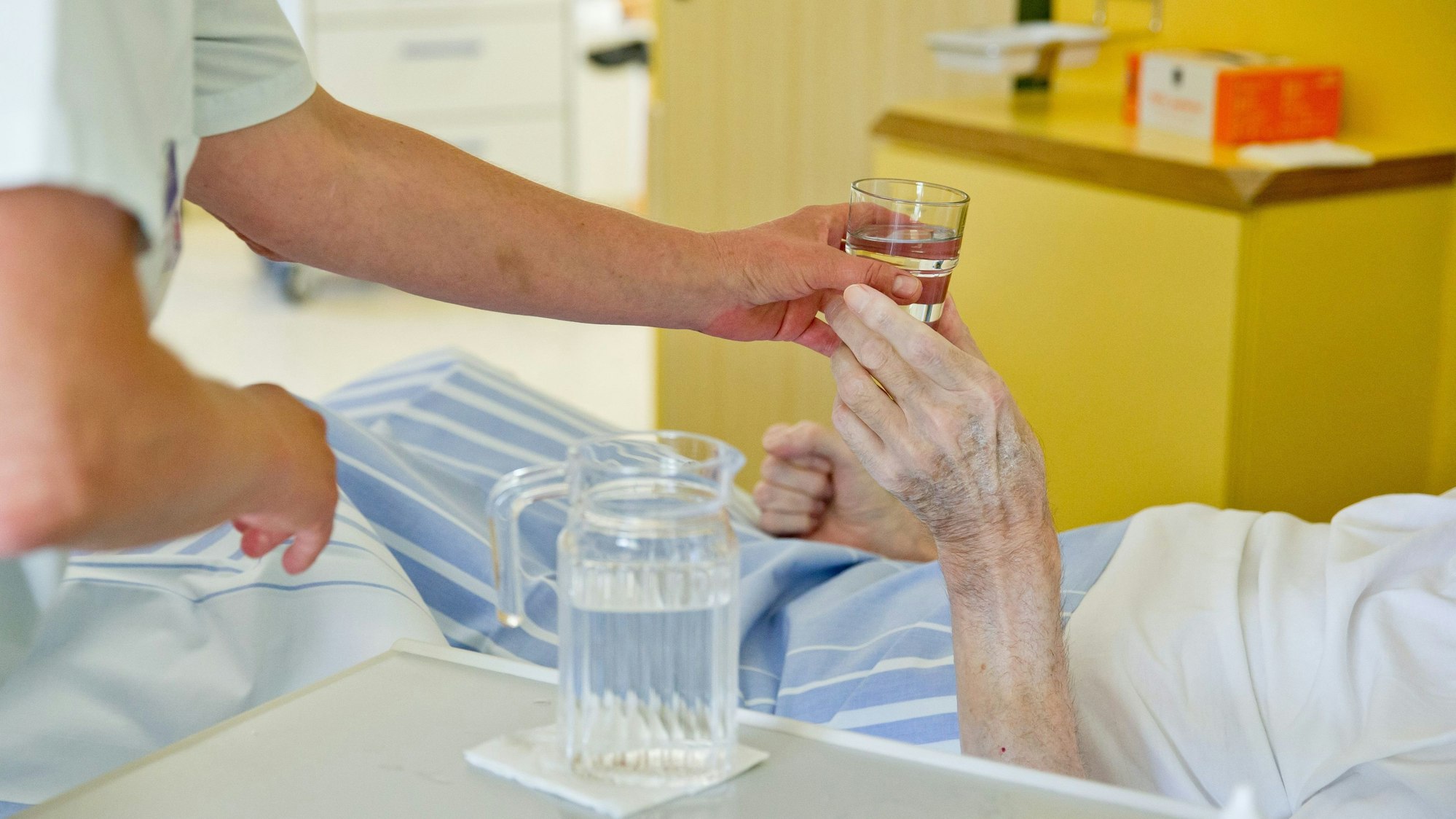 Eine Pflegerin reicht einer im Bett liegenden Seniorin ein Glas Wasser.