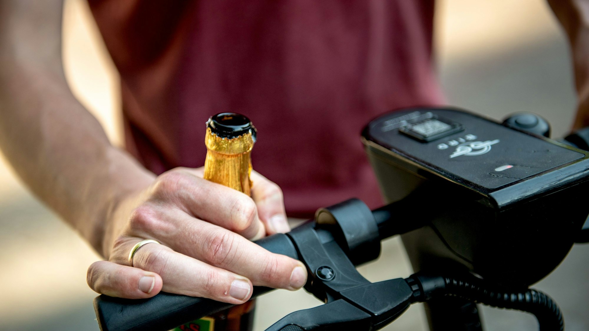 Das Foto zeigt eine Hand, die eine Bierflasche hält und das Lenkrad eines E-Scooters umfasst.