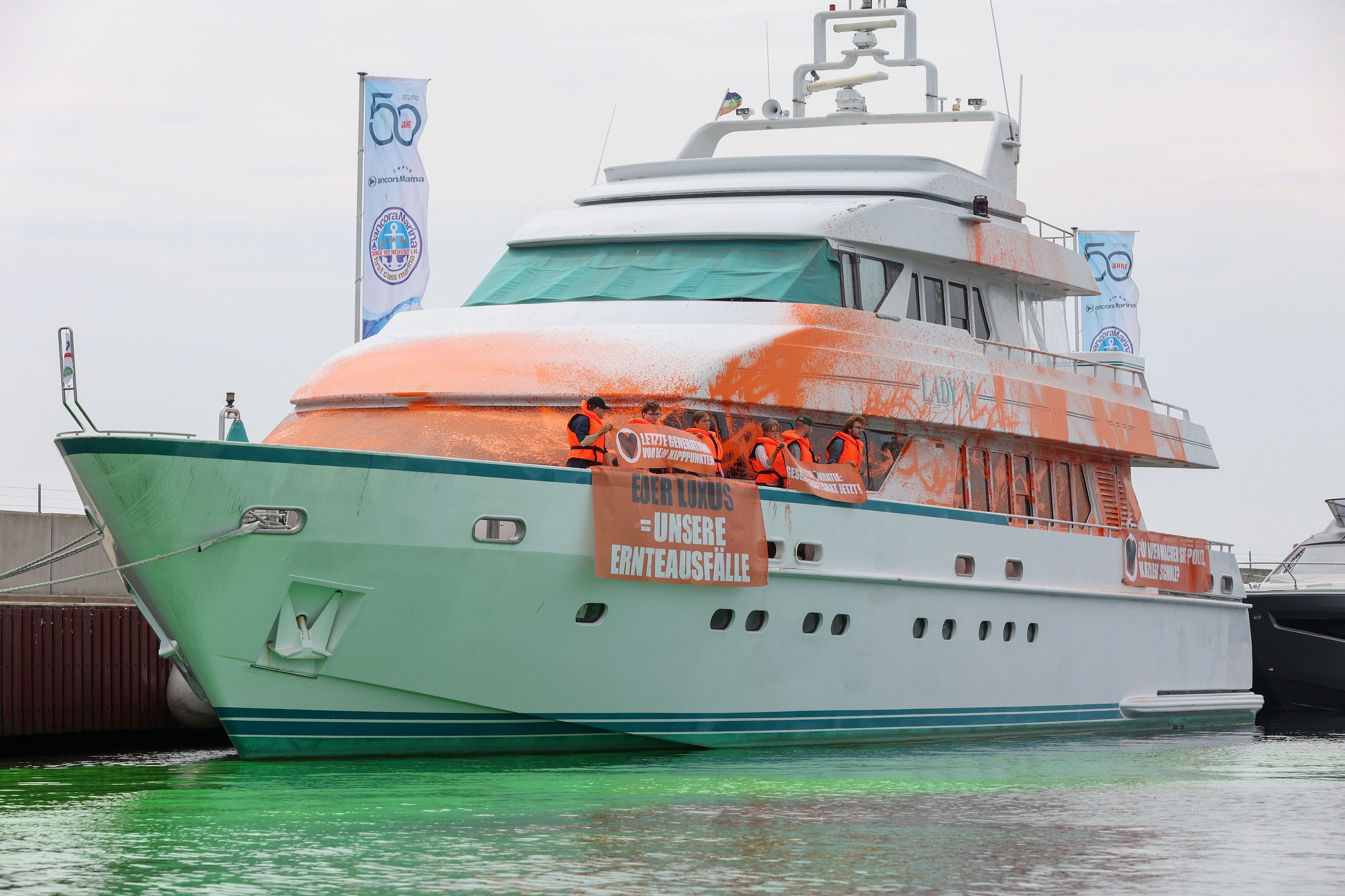 Teilnehmer einer Protestaktion stehen mit Transparenten auf einer Yacht im Hafen von Neustadt. Auf einem Transparent ist die Aufschrift Euer „Luxus = Unsere Ernteausfälle“ zu sehen.