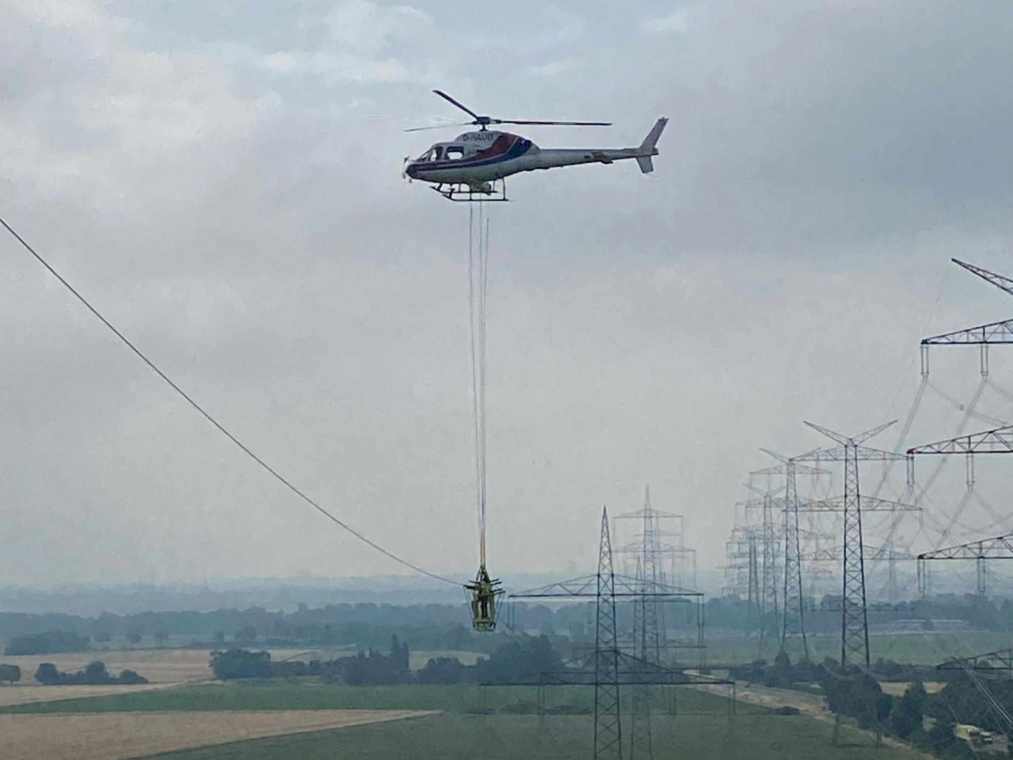 Das Foto zeigt einen Monteur am Seil unter einem Hubschrauber vor einer Kulisse aus Hochspannungsmasten.