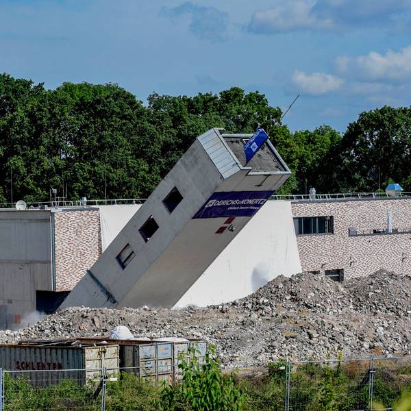 Das Foto zeigt einen umkippenden Turm vor einem Neubau.