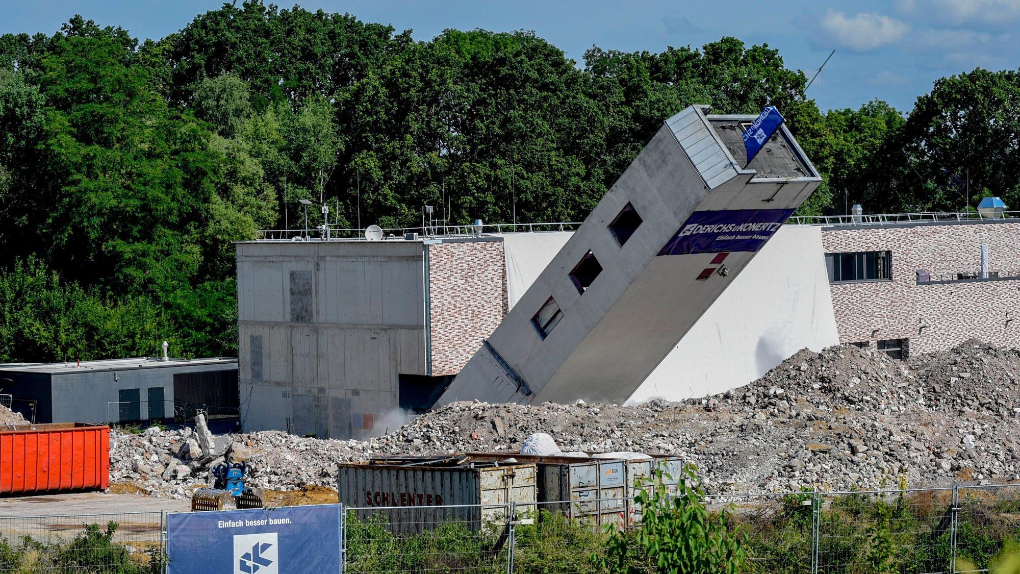 Das Foto zeigt einen umkippenden Turm vor einem Neubau.