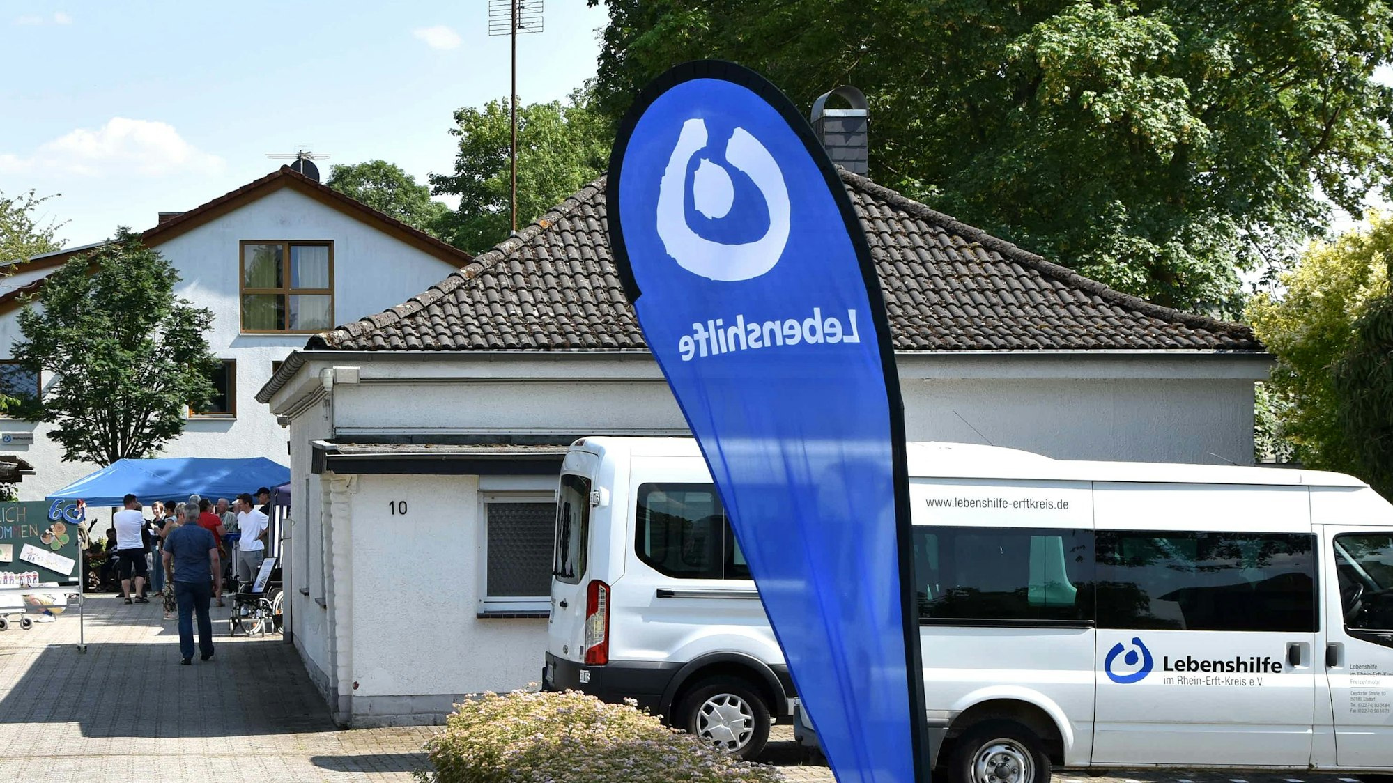 Das Bild zeigt das Gelände der Lebenshilfe an der Desdorfer Straße, ein „Lebenshilfe“-Aufsteller und Autos mit „Lebenshilfe“-Aufschrift sind im Vordergrund zu sehen.