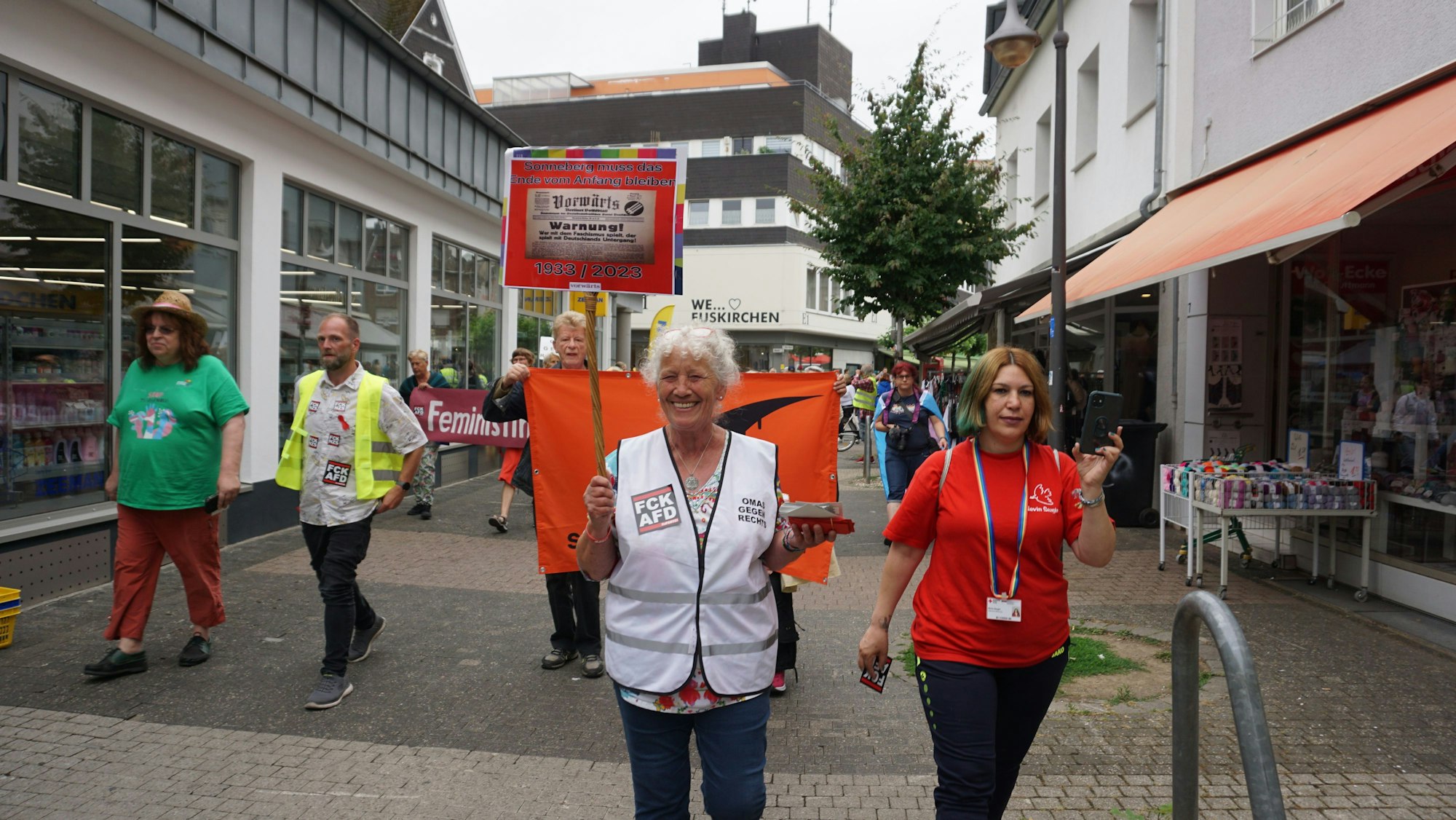 Eine Frau trägt eine weiße Sicherheitsweste. Sie hält ein Schild hoch.