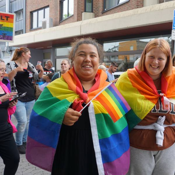 Zwei junge Frauen haben sich Regenbogenfahnen umgehangen. Eine von ihnen schwenkt zusätzlich eine Regenbogenflagge.