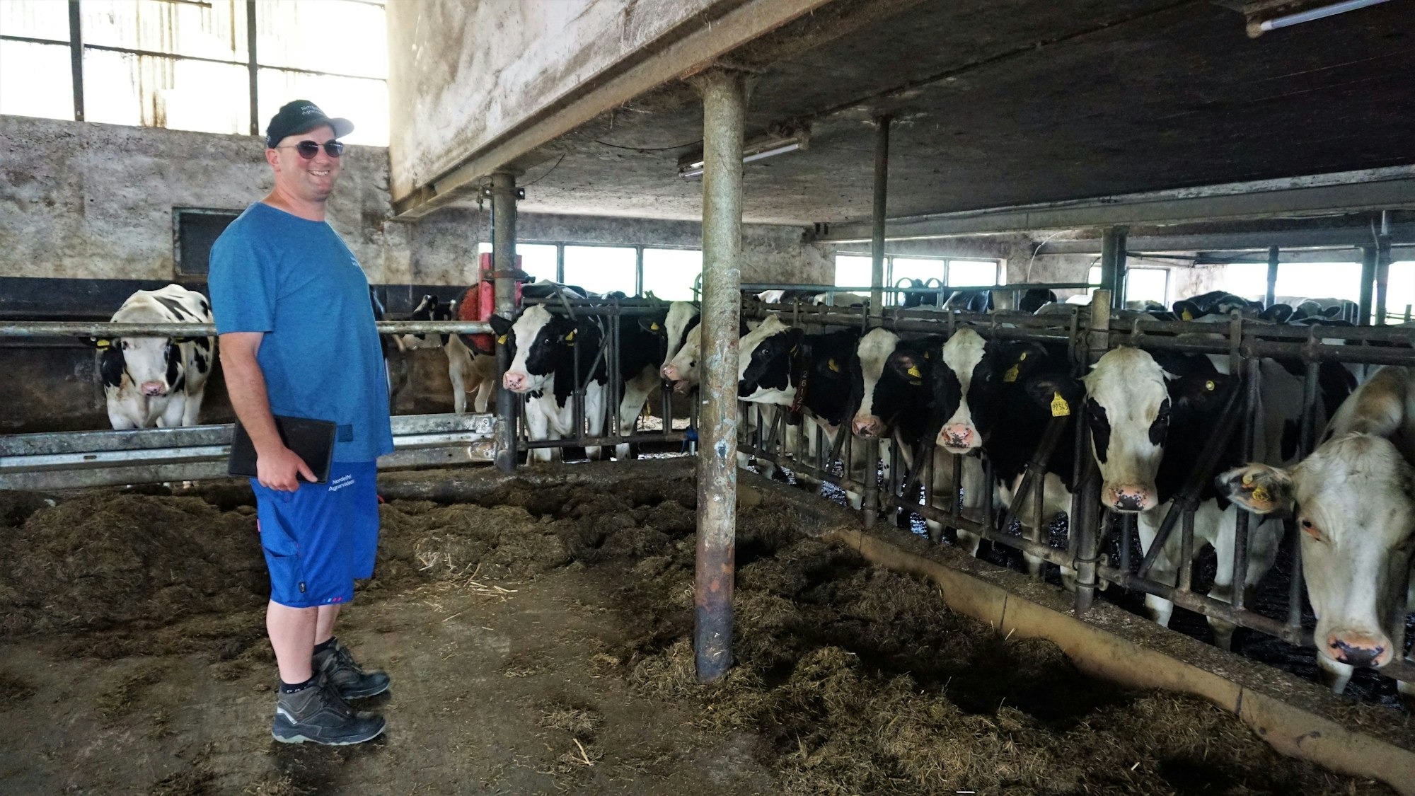 Sebastian Bützler steht im Kuhstall und hält sein Tablet in der Hand. Im Hintergrund befinden sich rund ein Dutzend Kühe.