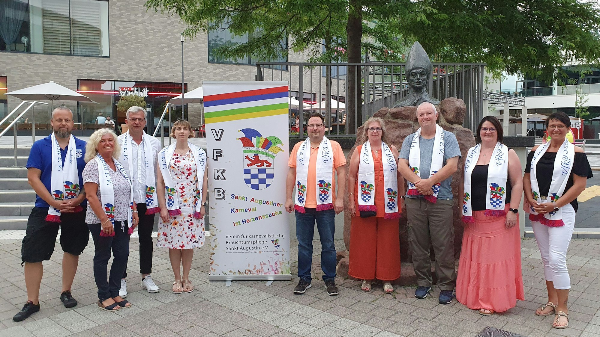 Neben einem Banner stehen die neun Menschen aus dem Vorstand. Sie alle tragen einen weißen Schal des Vereins.