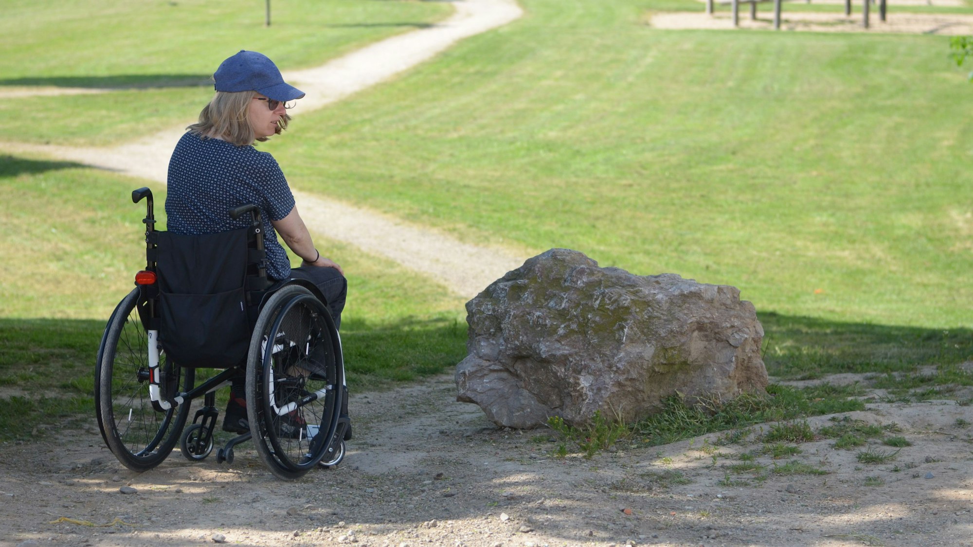 Die Rollstuhlfahrerin auf einem Weg vor einer Rasenfläche und einem großen Stein.