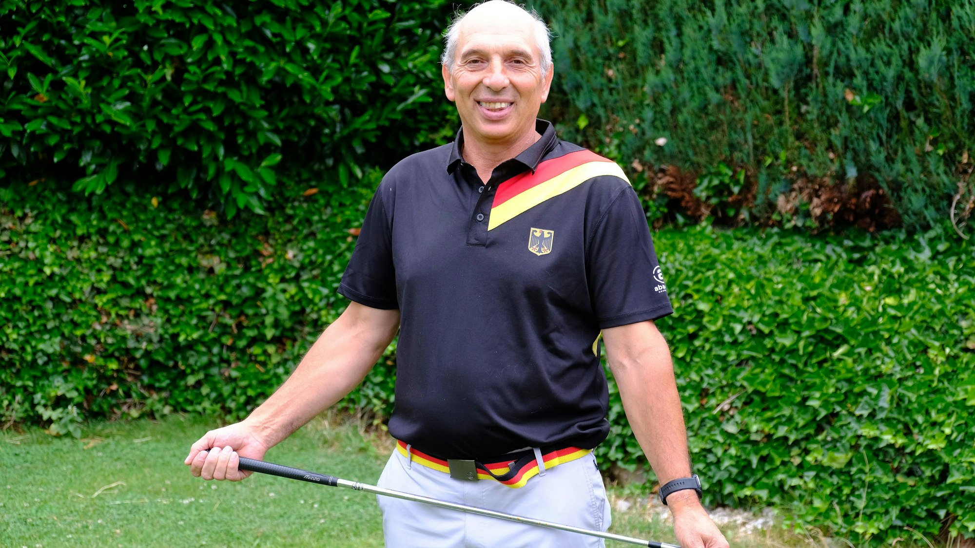 Der Golfspieler trägt ein Poloshirt mit Bundesadler und den deutschen Farben und hält einen Golfschläger in den Händen.