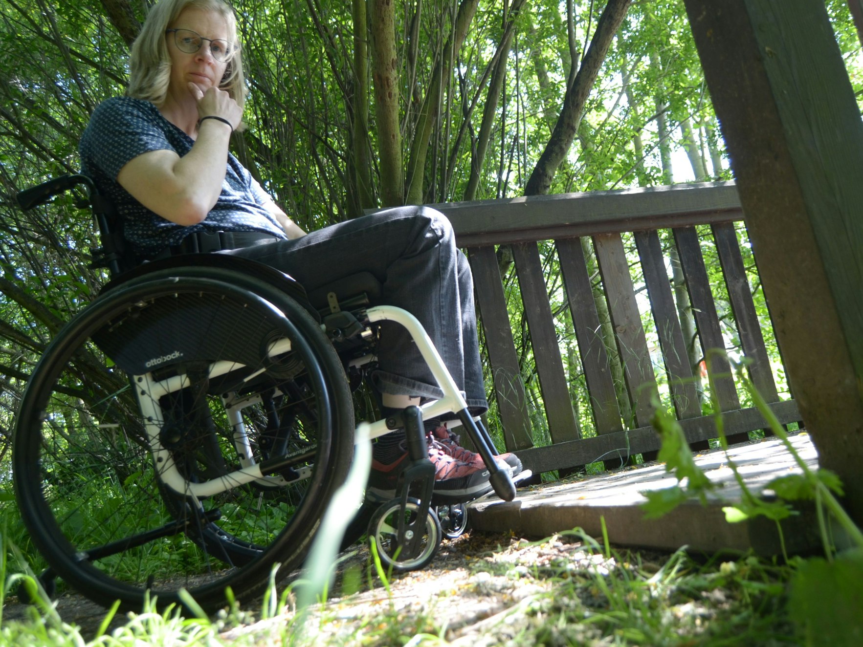 Eine Rollstuhlfahrerin vor der Stufe zu einer hölzernen Brücke.