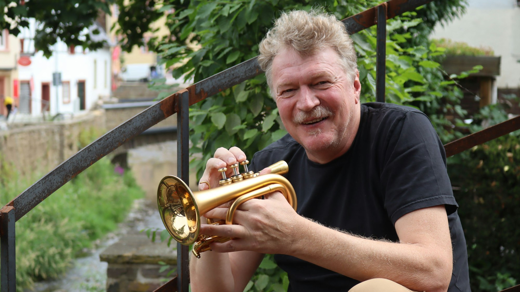 Hans Peter Salentin sitzt im Café Marielle in Bad Münstereifel auf einer rostigen Treppe, in seiner Hand hält er eine Mini-Trompete.