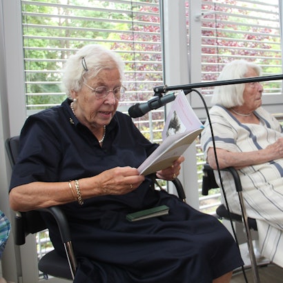 Drei ältere Damen sitzen nebeneinander, eine liest aus einem Buch, sie spricht in ein Mikrofon.