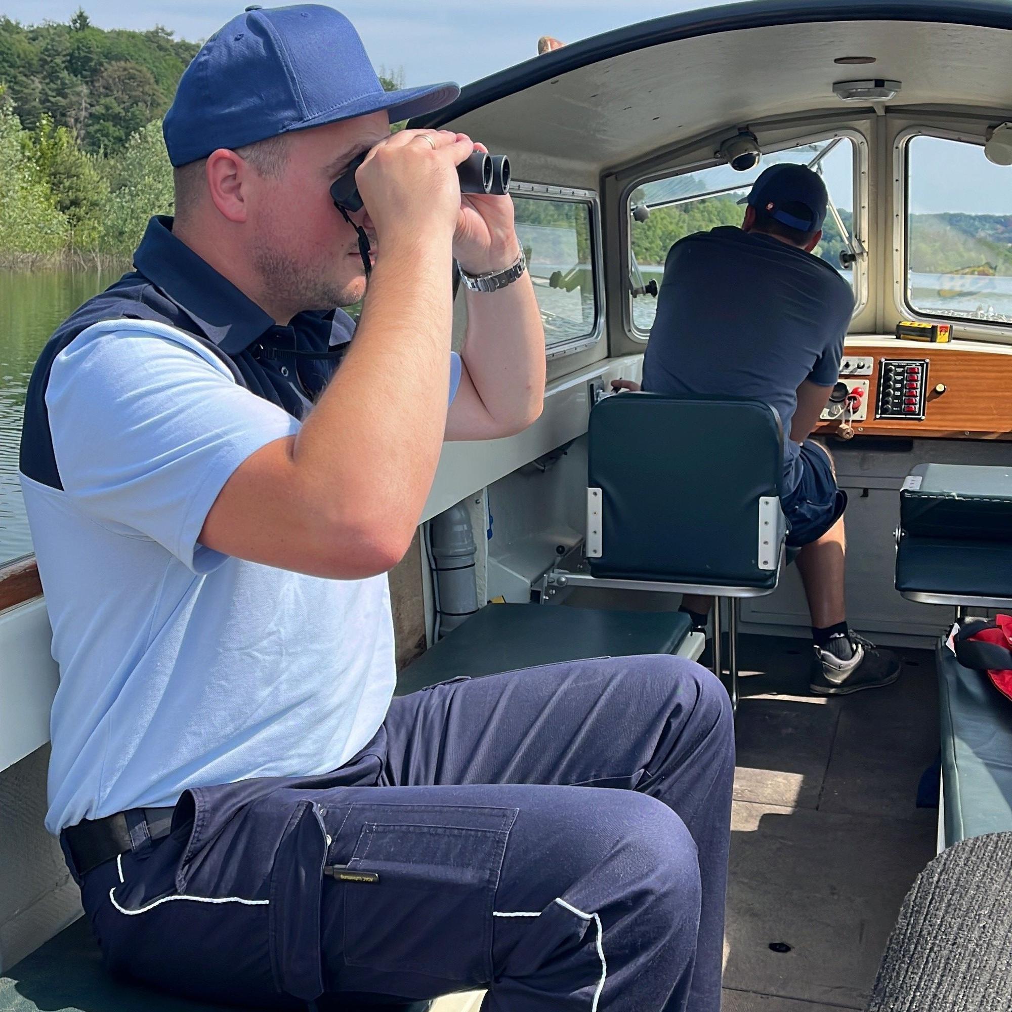 Ein uniformierter Wachmann beobachtet vom Elektroboot aus das Ufer der Talsperre durch ein Fernglas.