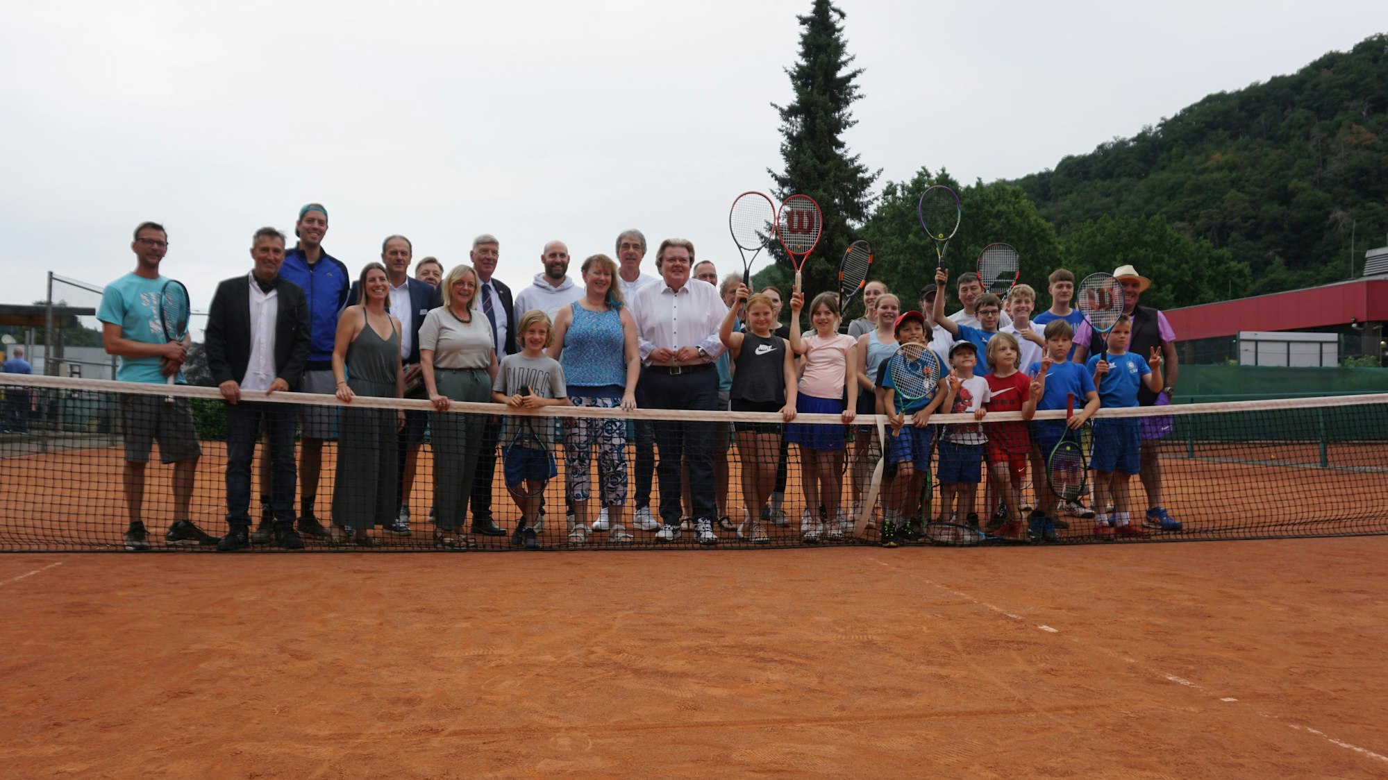 Eine Gruppe aus Kindern und Erwachsenen steht auf dem Tennisplatz. Die Kinder halten ihre Tennisschläger in die Luft.