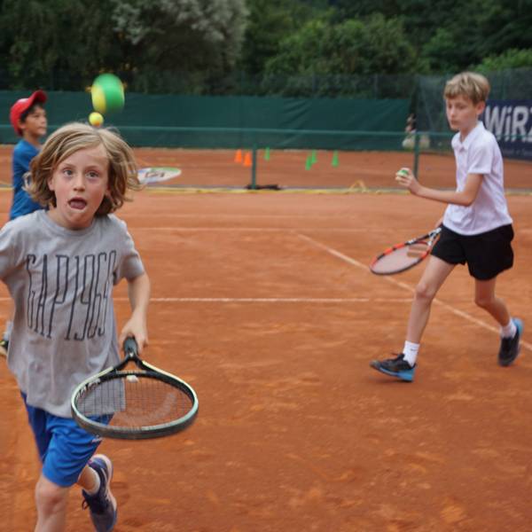 Ein Junge läuft mit einem Schläger über den Tennisplatz. Ein Tennisball fliegt durch die Luft. Der Junge hat einen konzentrierten Gesichtsausdruck.