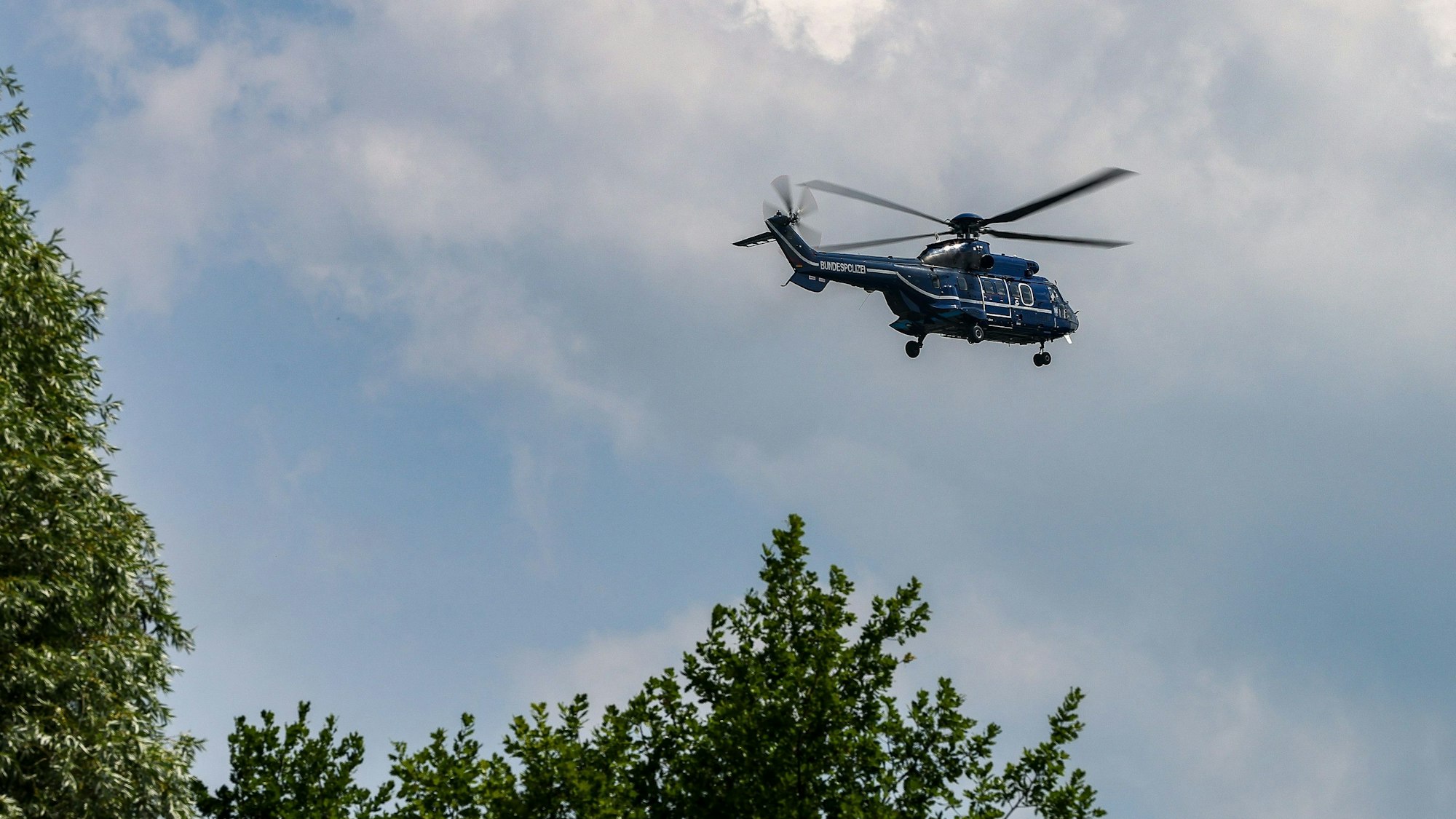 Ein dunkelblauer Hubschrauber fliegt vor leicht bewölktem Himmel über Baumspitzen.