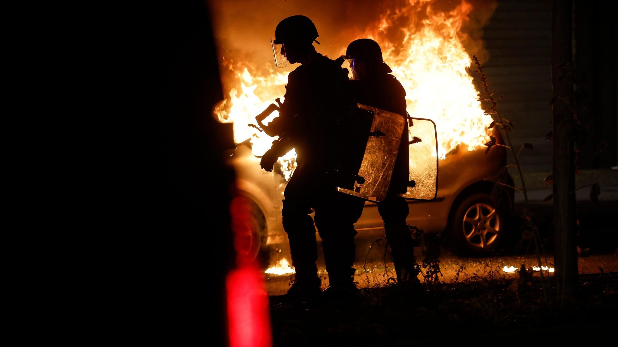 30.06.2023, Frankreich, Straßburg: Bereitschaftspolizisten stehen in der Nähe eines brennenden Autos im Viertel La Meinau. Mit Einschränkungen des öffentlichen Lebens will die Regierung in Frankreich die schweren Krawalle nach dem Tod eines Jugendlichen durch einen Polizeischuss eindämmen. Foto: Jean-Francois Badias/AP/dpa +++ dpa-Bildfunk +++