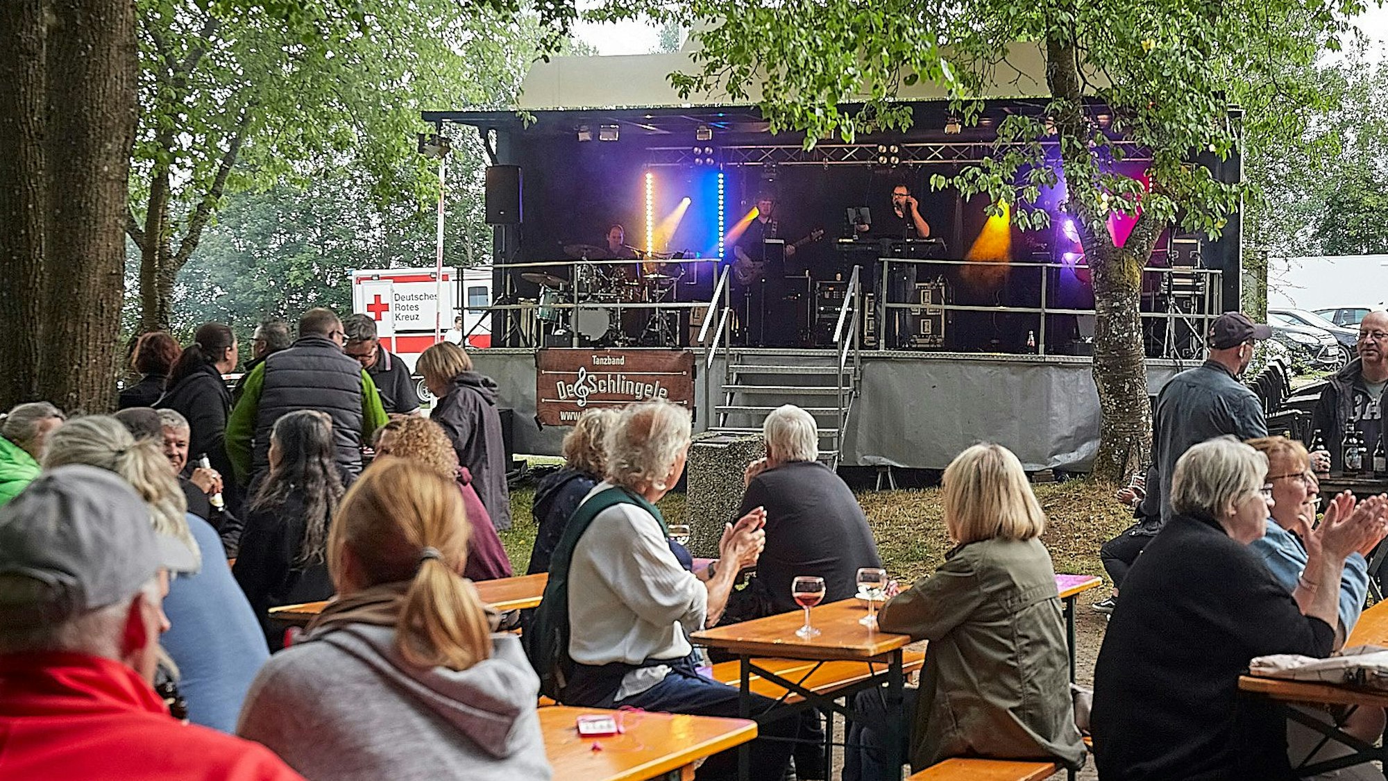 Besucher sitzen an Bierbänken. Auf einer Bühne spielt die Band „De Schlingele“.