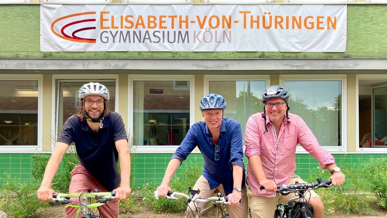 Drei Männer auf Fahrrädern stehen vor einem Schulgebäude