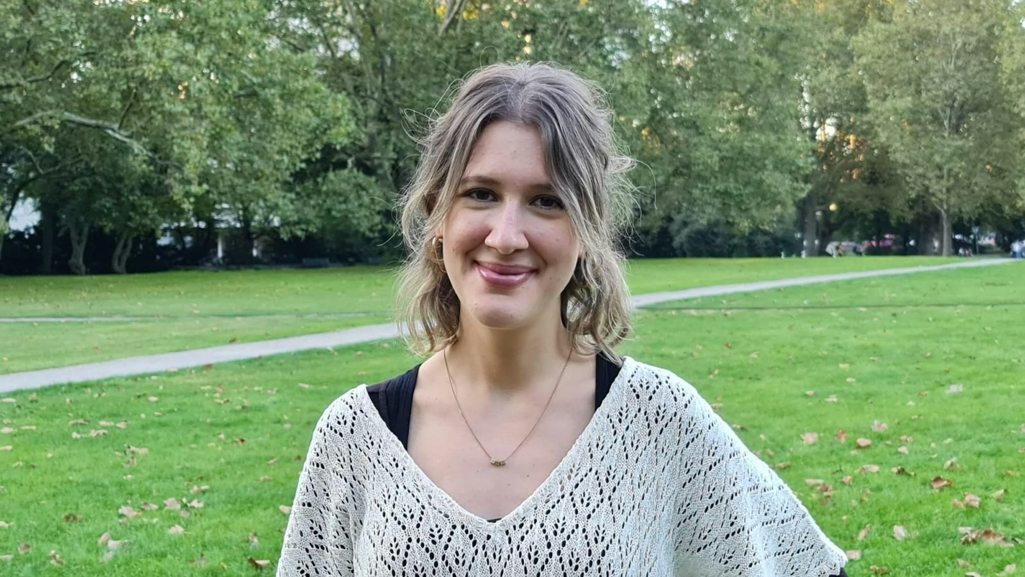 Eine junge Frau mit blondem kinnlangem Haar steht in einem Park.