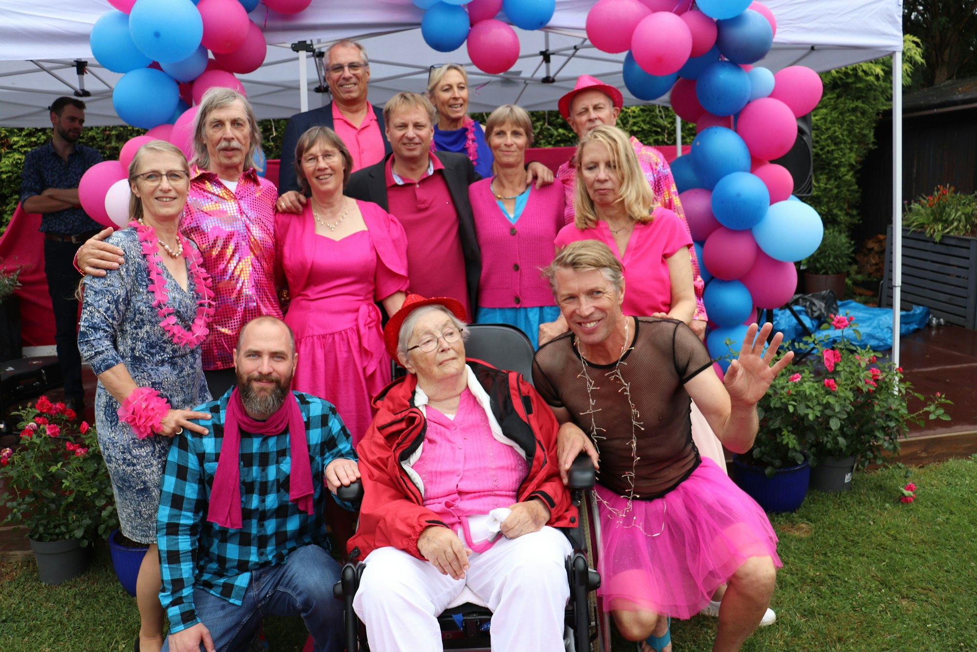 Zwölf Personen in Blau und Pink sind zu sehen. Sie umrahmen eine ältere Dame vorne in der Mitte.