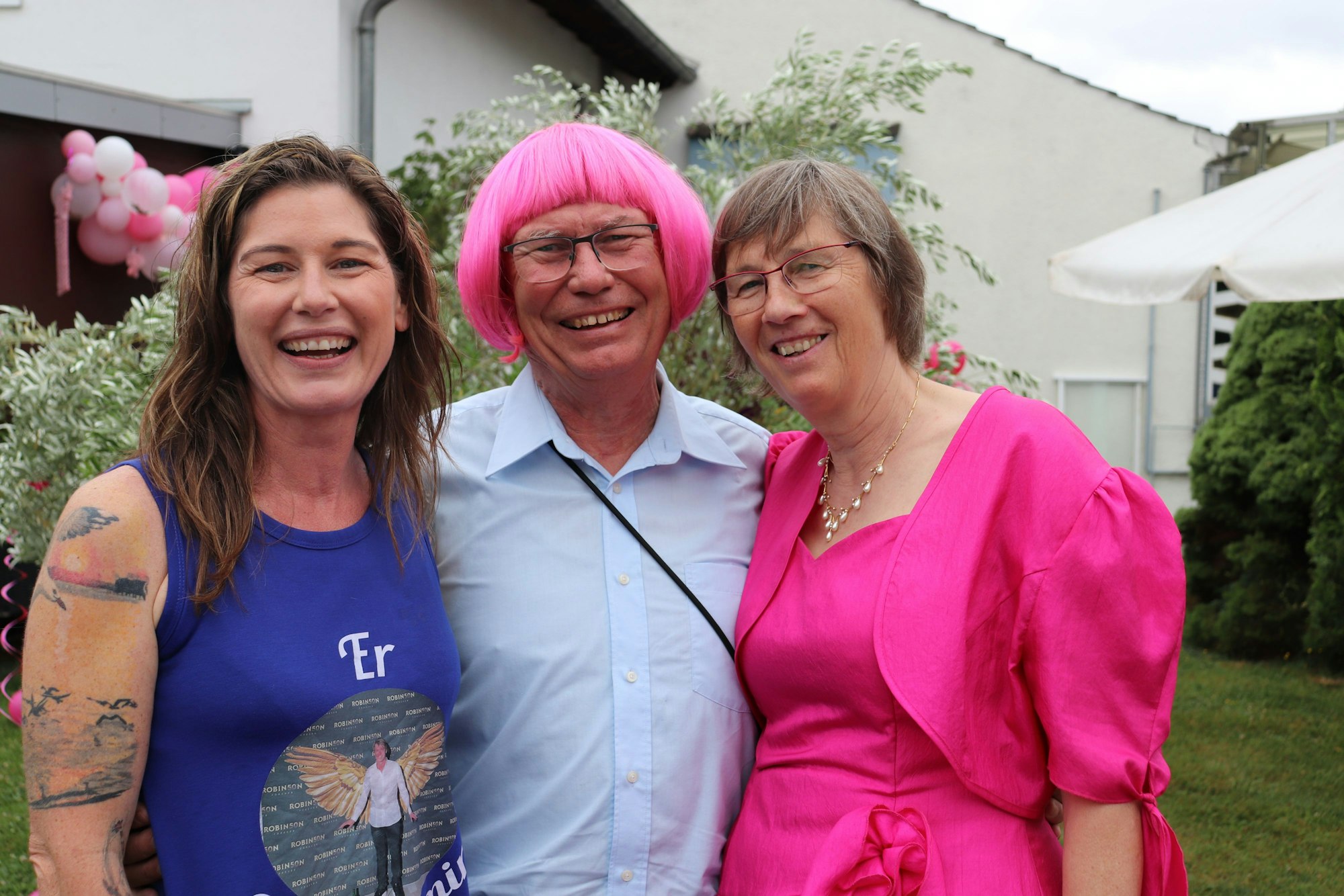 Zwei Frauen umrahmen einen Mann mit pinker Perücke.
