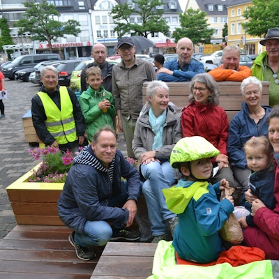 Erwachsene und Kinder in bunten Regenjacken sitzen auf Bänken und Blumenkästen aus Holz. Alle lachen.