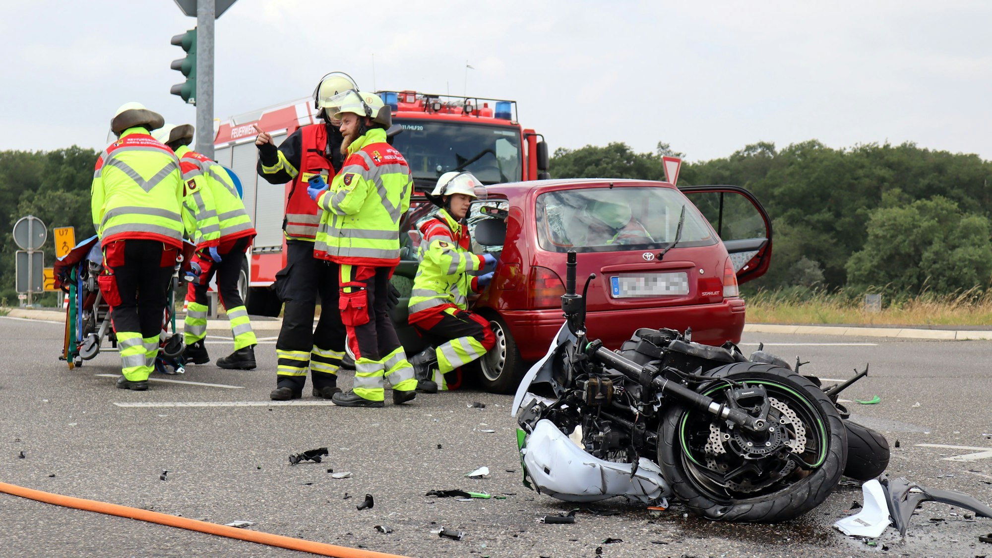 Ein schwer beschädigtes Motorrad liegt auf der Fahrbahn, dahinter stehen das an dem Unfall beteiligte Auto und ein Feuerwehrfahrzeug. Der Rettungsdienst kümmert sich um Verletzte.