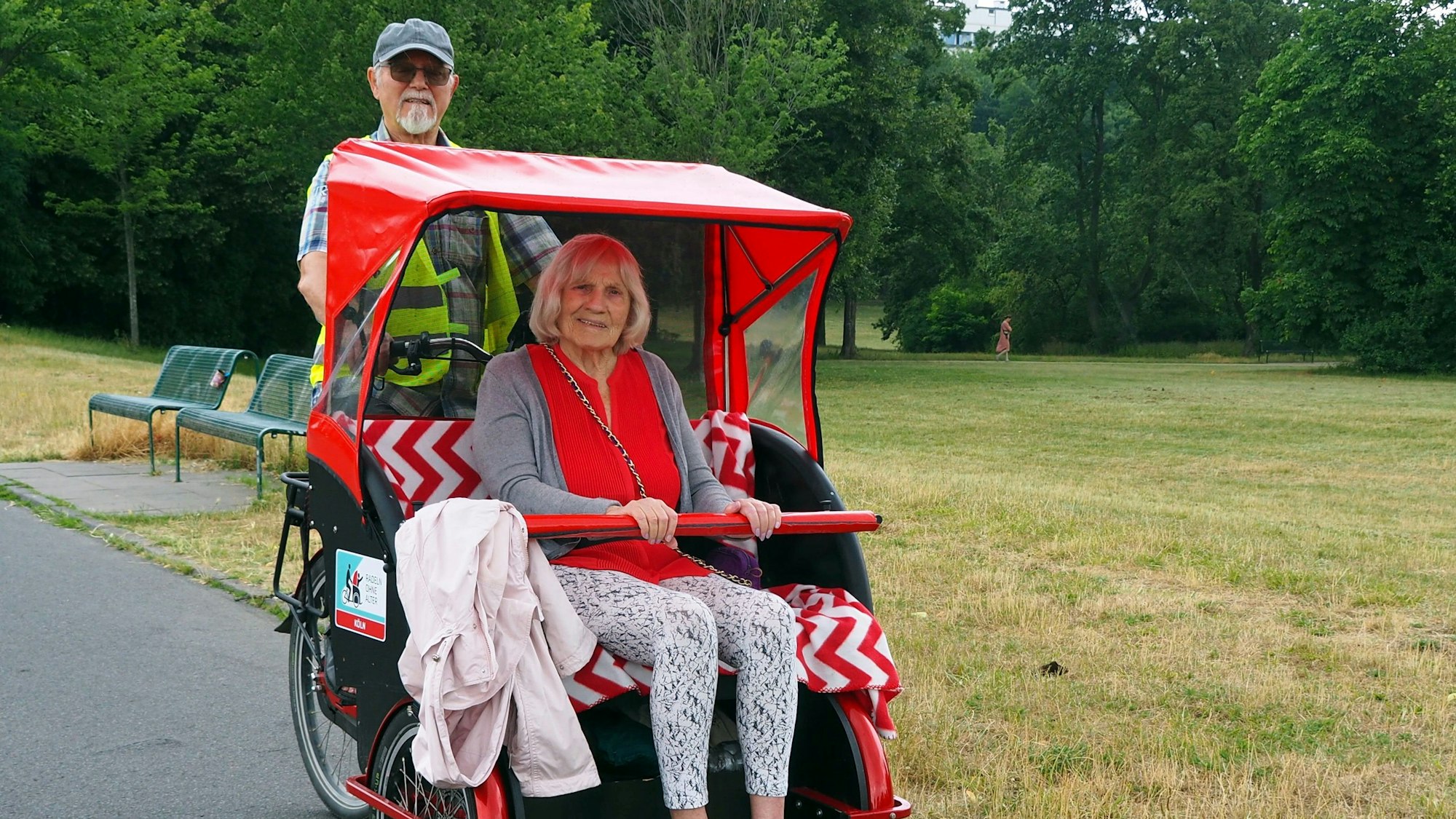 Eine ältere Dame sitzt in einer Rikscha, die ein Herr mit Bart und Schirmkappe fährt.