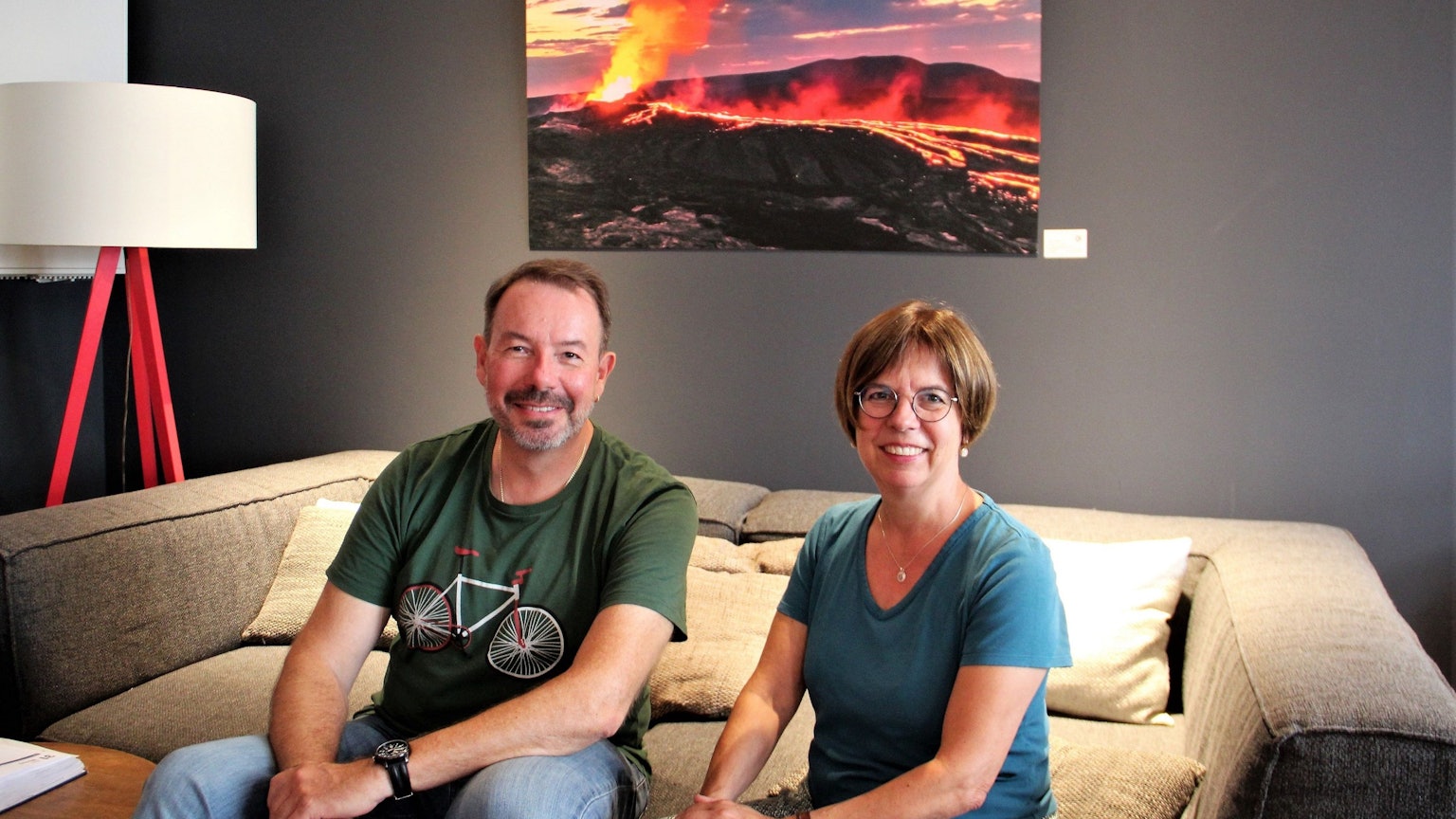 Ein Mann mit Bart und eine Frau mit Pagenkopf und Brille sitzen auf einem Sofa, hinter sich ist ein Bild von einem Vulkan-Ausbruch zu sehen.