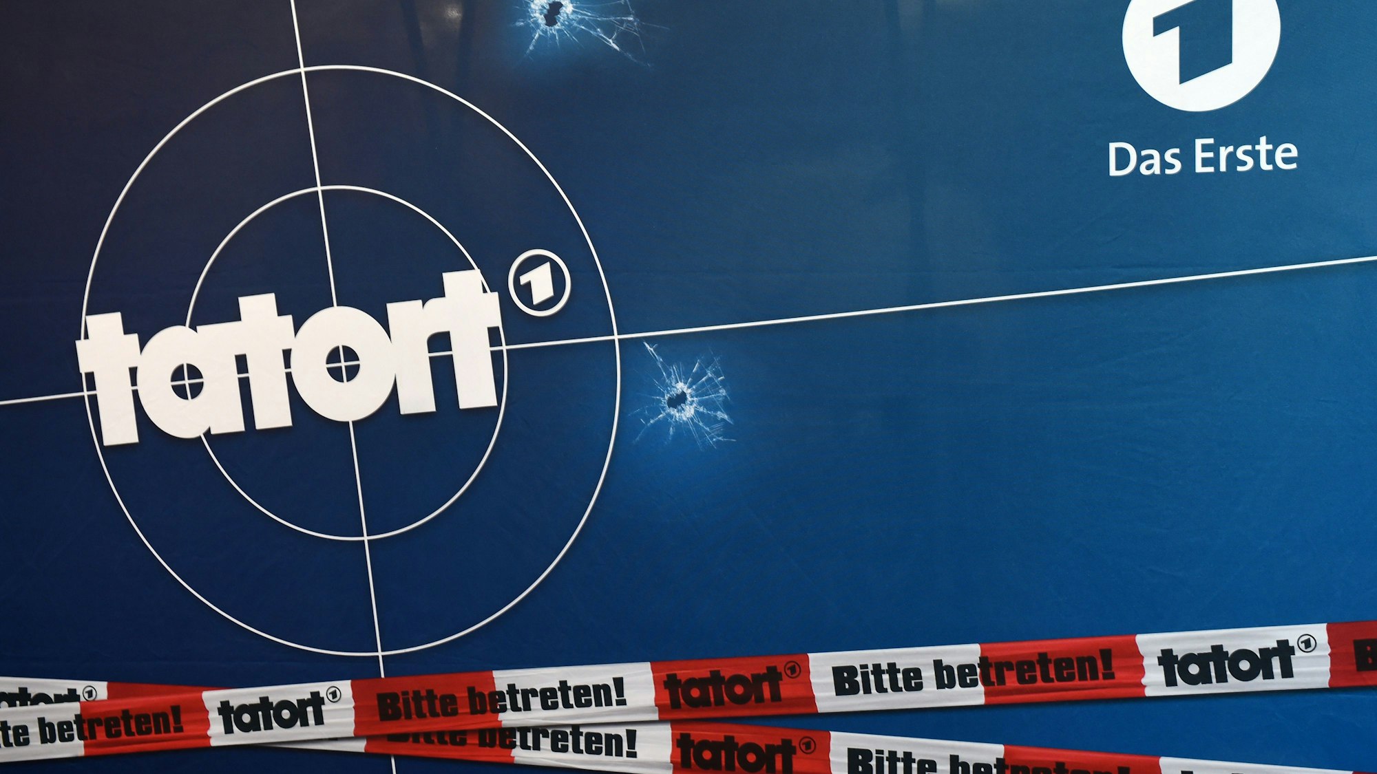 Auf einer Fotorückwand sind das Logo der ARD-Fernsehreihe „Tatort“ zu sehen, zudem ein rot-weißes Absperrband.