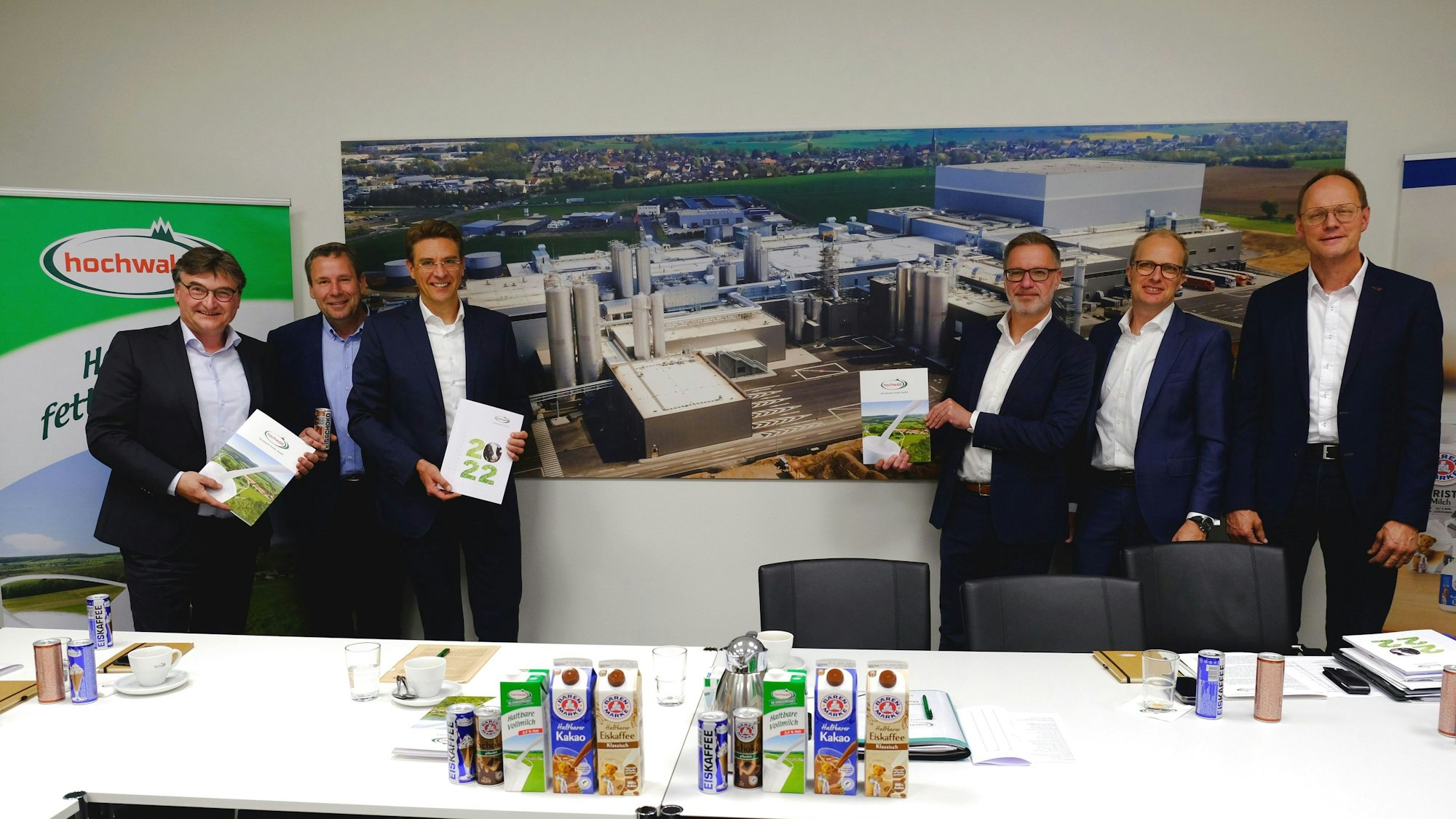 Mehrere Männer in Anzügen stehen vor einem großen Wandbild, das die Molkerei in Obergartzem zeigt.