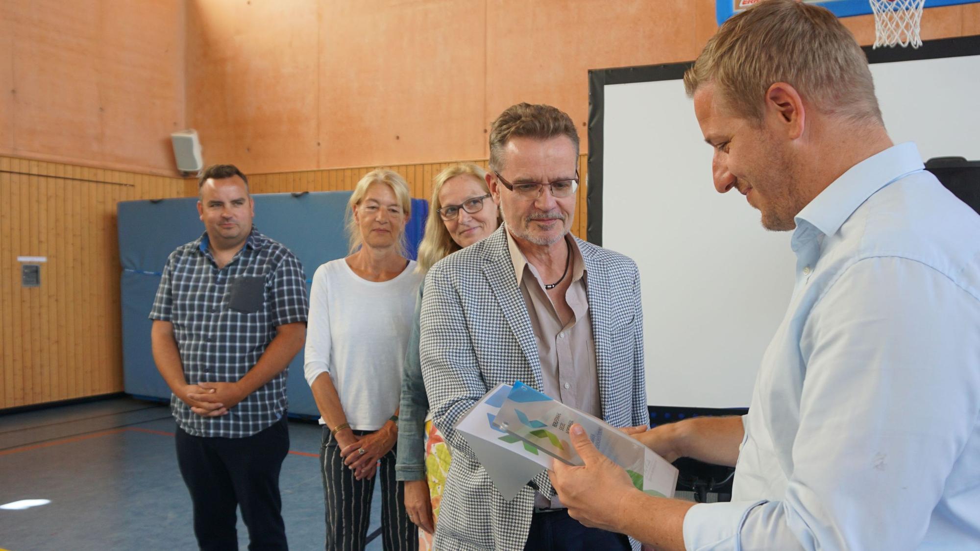 Landrat Markus Ramers und Vertreter der ausgezeichneten Schulen stehen in einer Turnhalle. Ramers übergibt die Plakette und die Urkunde.