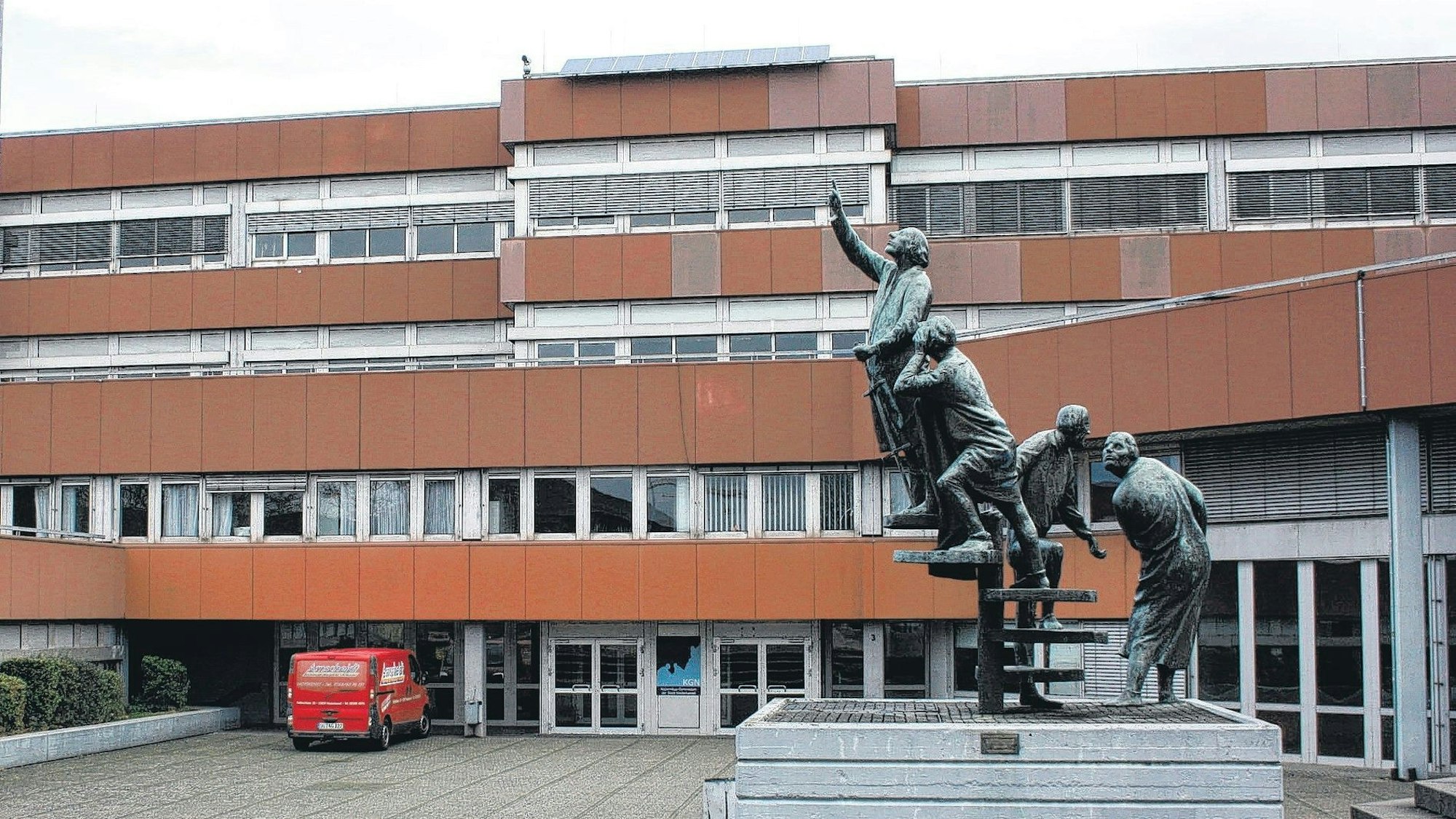 Ein Schulgebäude mit rostroter Fassadenverkleidung. im Vordergrund steht eine Skulptur, die vier Figuren darstellt.