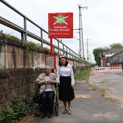 Ein Mann sitzt auf seinem Rollator vor dem Schild „Haltestelle außer Betrieb“. Neben ihm steht seine Frau.