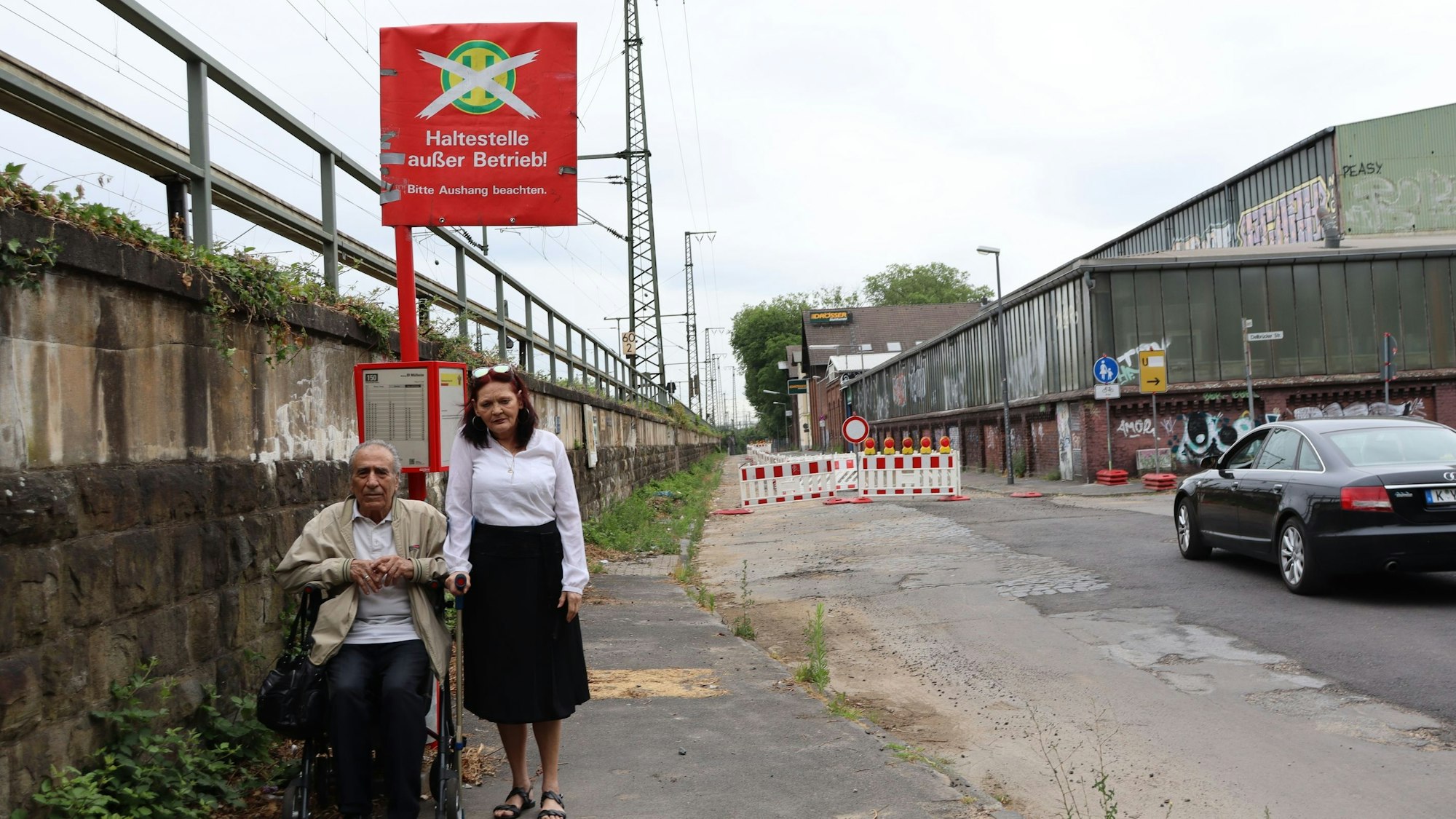 Ein Mann sitzt auf seinem Rollator vor dem Schild „Haltestelle außer Betrieb“. Neben ihm steht seine Frau.