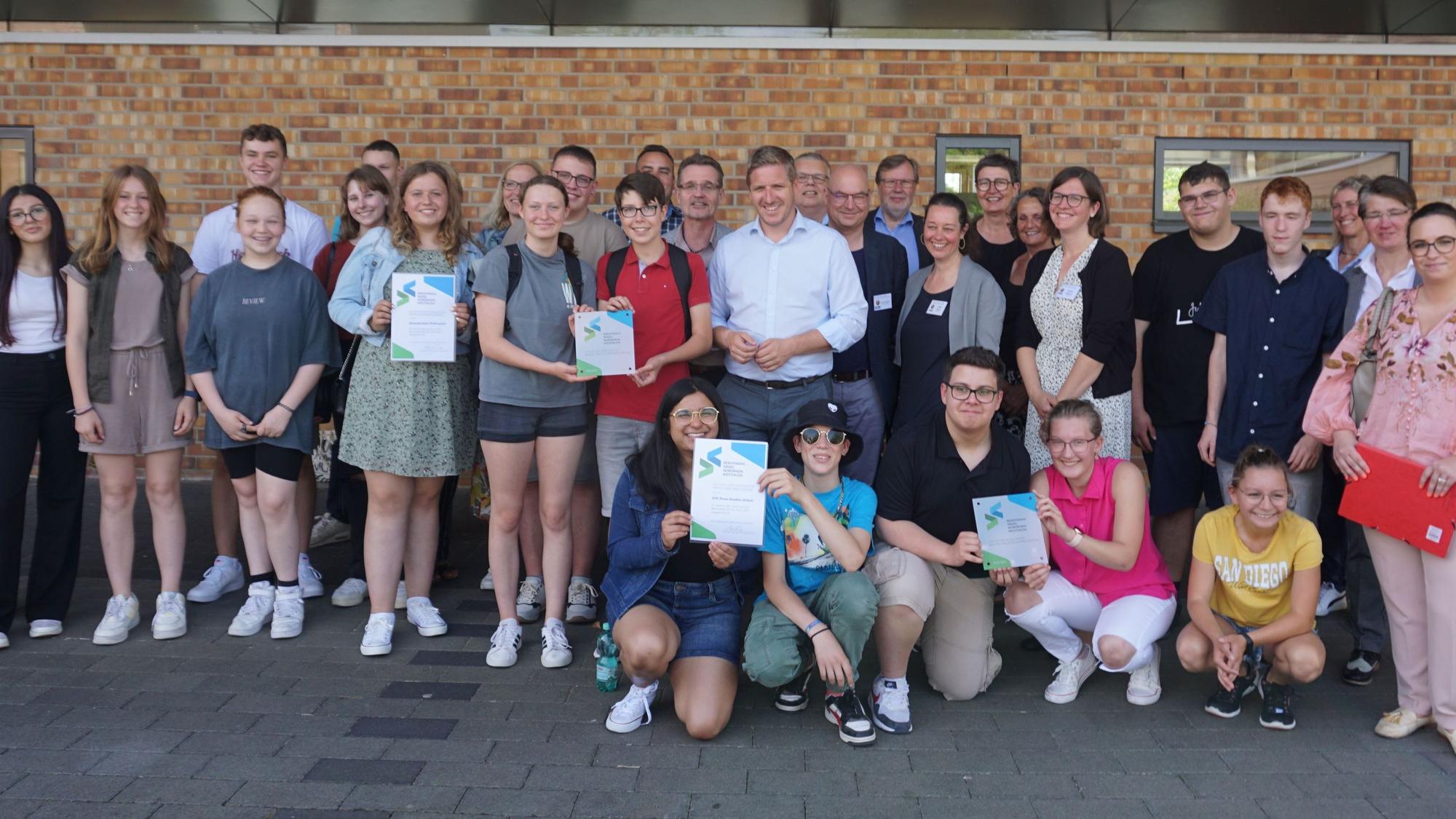 Schüler und Schulleiter der LVR-Irene-Sendler Schule und der Gesamtschule Weilerswist stehen vor einer Mauer und halten Urkunden hoch.