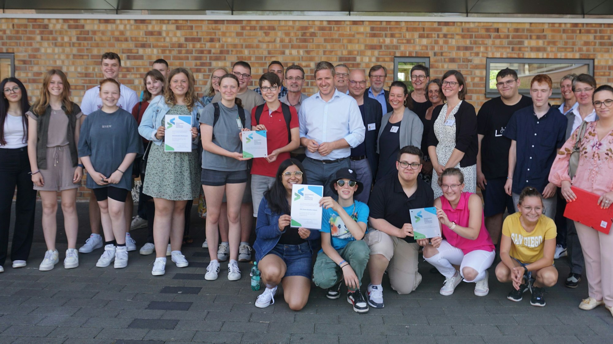 Schüler und Schulleiter der LVR-Irene-Sendler Schule und der Gesamtschule Weilerswist stehen vor einer Mauer und halten Urkunden hoch.