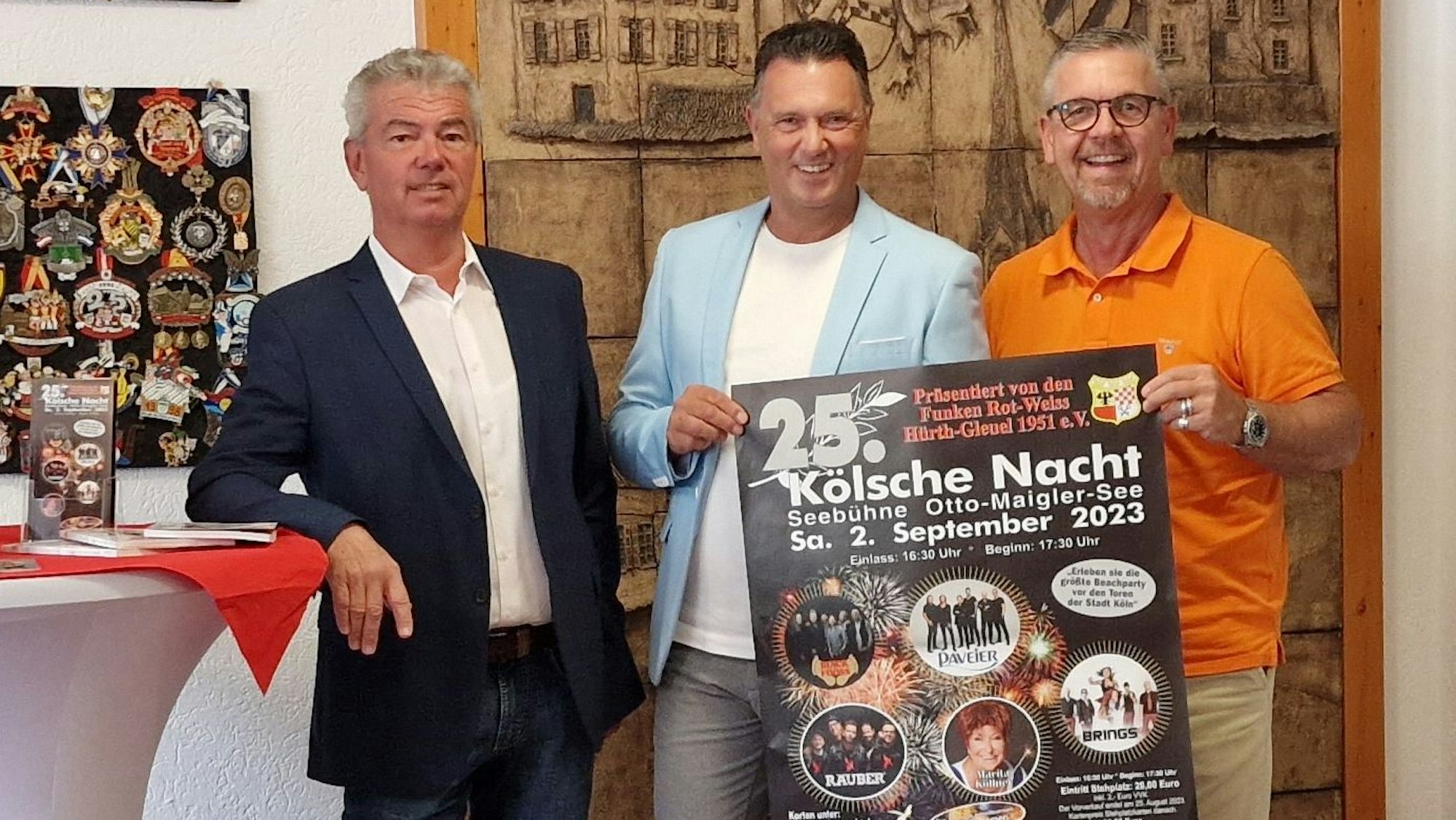 Drei Männer stehen vor einer Pinwand mit Karnevalsorden und präsentieren ein Plakat.
