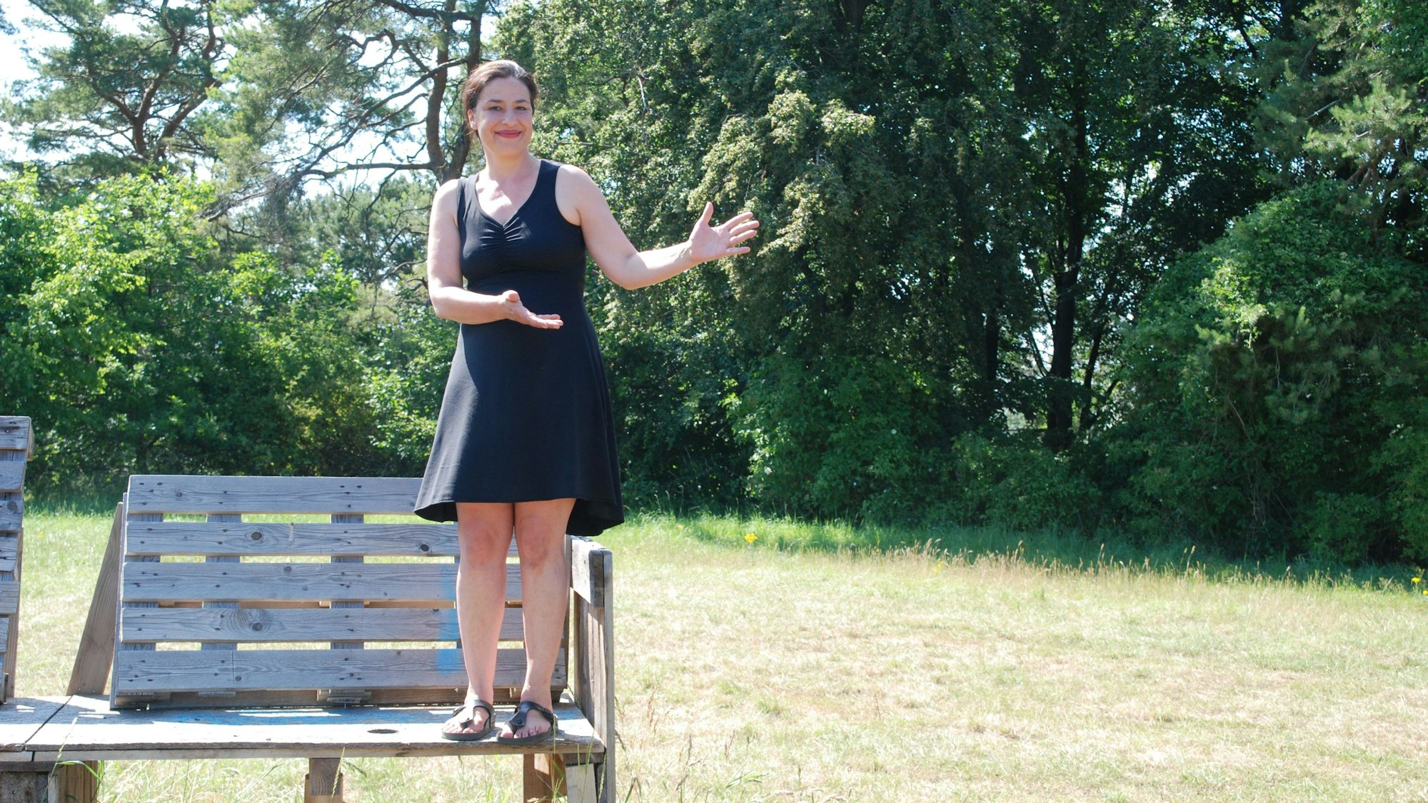 Kabarettistin Eva Eiselt steht auf einer Tribüne aus Euro-Paletten. Sie trägt ein schwarzes Sommerkleid und zeigt auf eine Wiese.